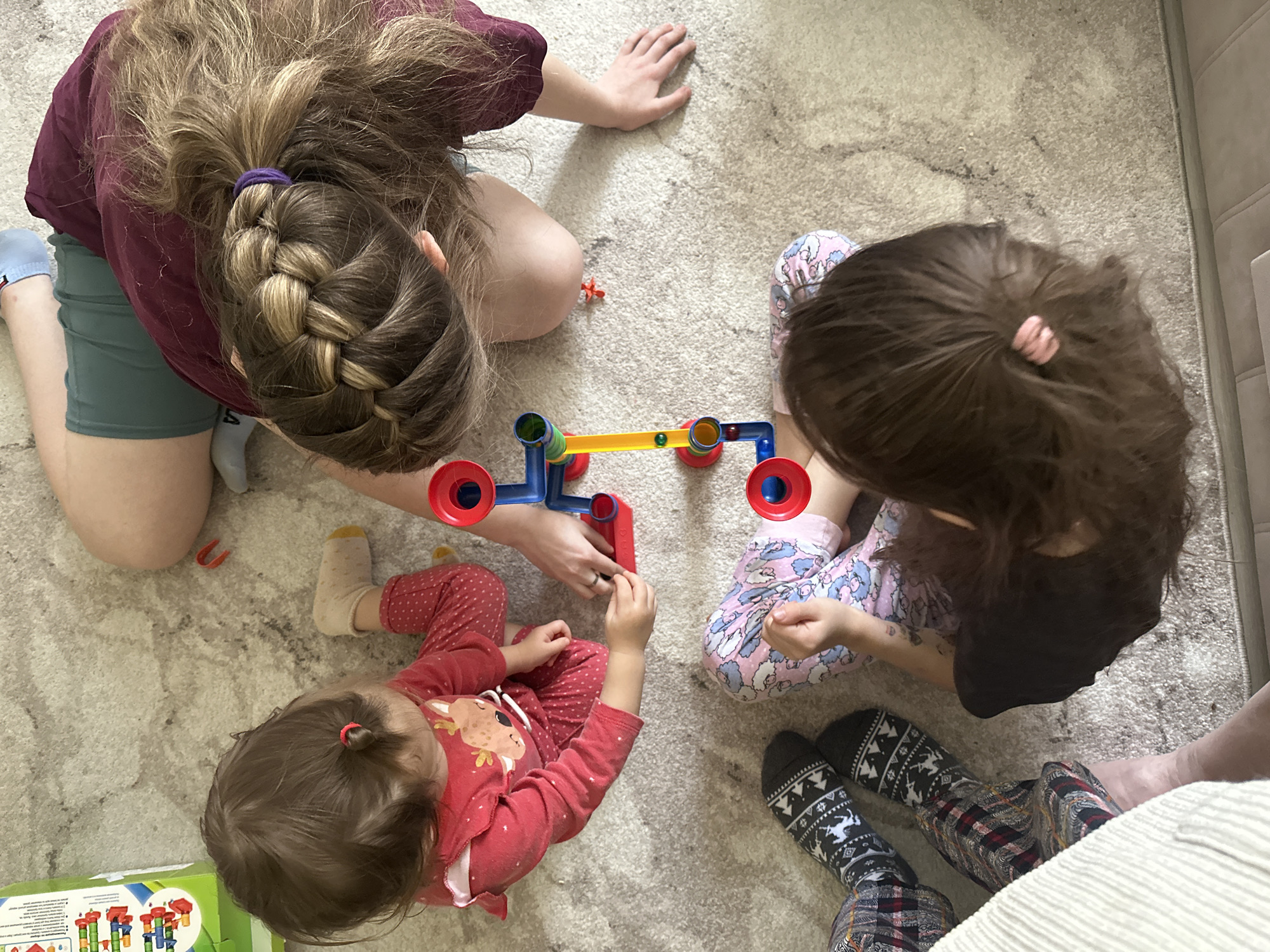 Four individuals, observed from an overhead perspective, are engaged in play with a toy marble run set constructed of blue, green, red, and yellow components, on a light-colored carpet. The individuals are positioned around the toy in a semi-circle.
One individual, possibly an adult, is wearing a maroon-colored shirt and green shorts, with blonde-streaked brown hair styled in a braid secured with a purple hair tie. A younger child, dressed in red pajamas adorned with a deer motif, is seated to the left of the adult. Another child is wearing pink and blue patterned pajama pants, while the fourth individual, potentially another adult, is wearing patterned socks and plaid pants.
The location is an interior space. The location in Uva, Russia cannot be determined from this image.