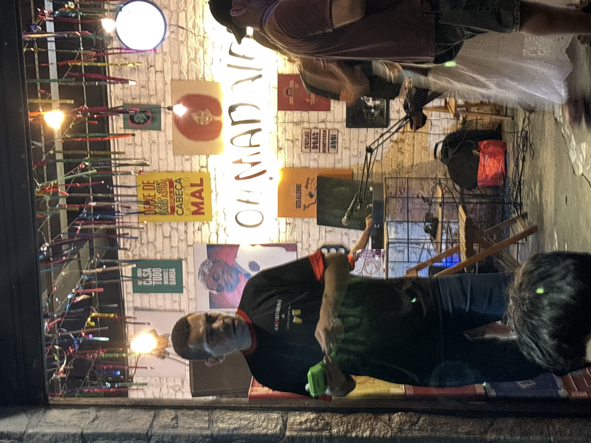 Here's a description of the scene:
The photo appears to have been taken through a glass window, possibly the front of a bar or pub in the Vila Madalena neighborhood in São Paulo, Brazil. The name of the establishment, "OH MAD AI," is visible in large, illuminated letters on the wall. The walls are decorated with various posters and artwork. Colorful decorations hang from the top of the window, obscuring some of the view.
Inside, several people are present. A man in a black shirt with red accents and the "CONVADALENA" logo is in the foreground, holding a green bottle. Another person, wearing a dark shirt, stands in the background. The establishment has a casual, informal atmosphere, with assorted items like chairs, microphone, bags, and other items scattered around.