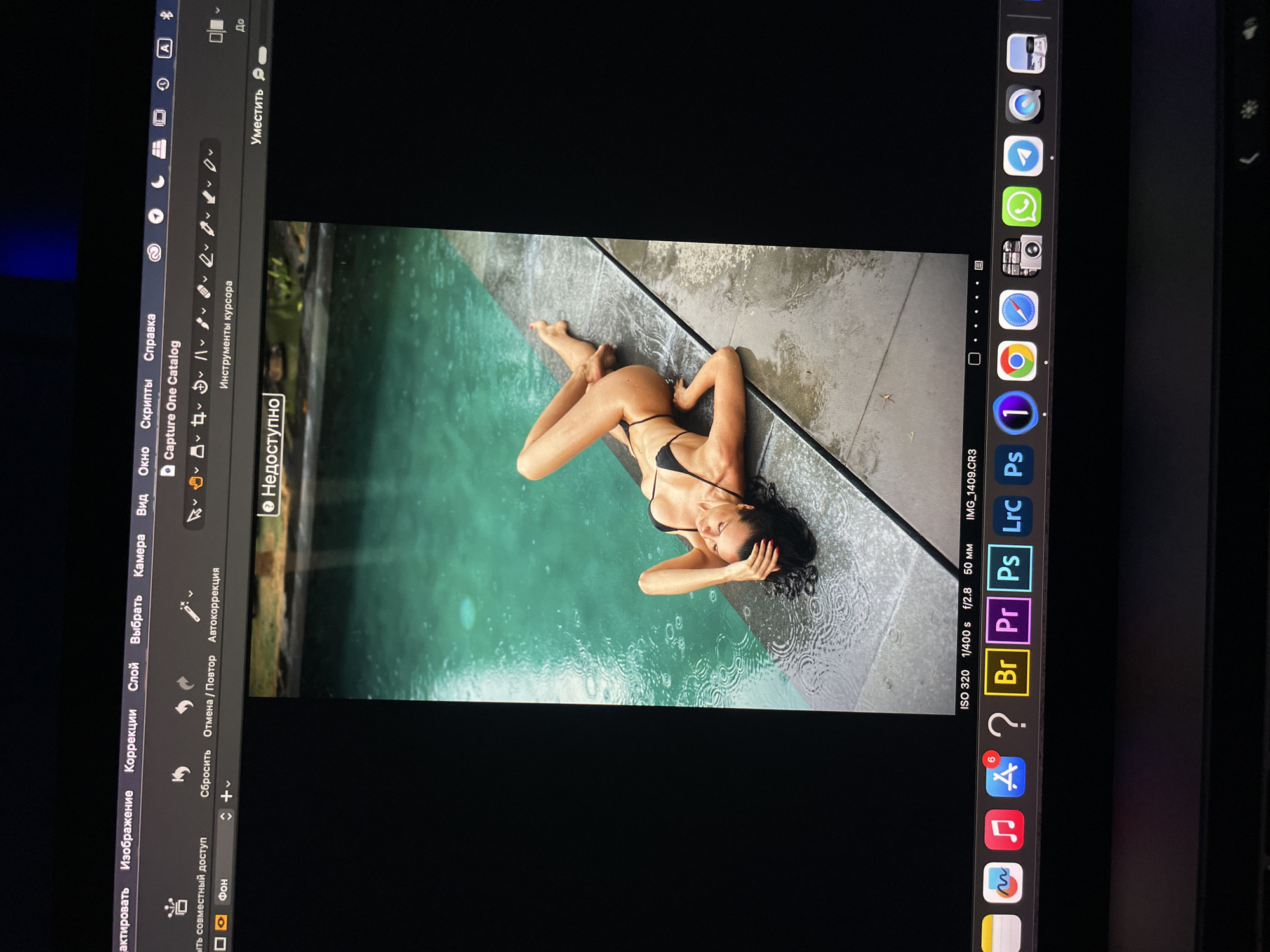 The image is a photograph of a woman in a black bikini lying on the edge of a pool. It appears to be raining, with raindrops visible on the surface of the water and the surrounding stone. The woman is partially submerged in the water. The photograph is open in Capture One editing software on a screen. The location is unknown.