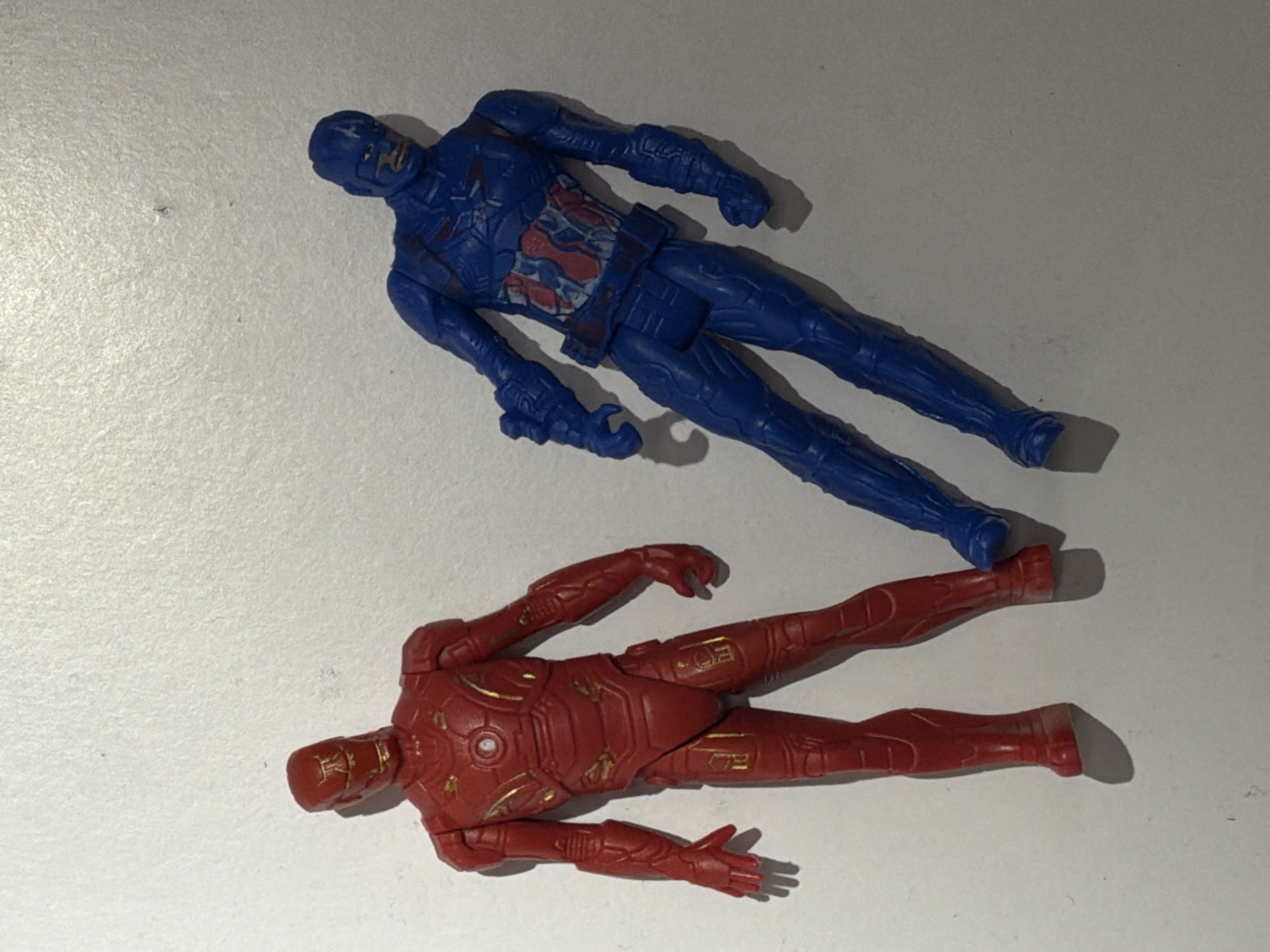 Two plastic action figures, one blue and one red, are depicted lying horizontally on a textured, light-colored surface in an indoor setting. The figures are positioned diagonally across the frame, with the blue figure above and slightly to the right of the red figure.
The upper figure is blue, resembling Captain America. It features molded details consistent with a superhero costume, including a segmented design, red and white markings on the chest area that suggest a star emblem and stripes, and a masked head. The figure shows some signs of wear, particularly on the chest and head, where the details appear slightly faded or smudged.
The lower figure is predominantly red with intricate gold-painted accents, characteristic of Iron Man's armor. It displays detailed panel lines, a circular indent on the chest, and gold markings on the limbs and torso. This figure also exhibits minor wear, with some of the gold paint appearing slightly chipped or rubbed off in places.
Both figures are presented in a static pose, implying no active event is taking place. They are simply resting on the surface, which appears to be a light grey or off-white material with a subtle grainy texture. Soft shadows are cast beneath the figures, indicating illumination from an overhead light source. There is no visible text or identifiable background elements to suggest a specific location within Sofiivska Borshchahivka, Ukraine, beyond the implied indoor environment. The image captures these superhero toy figures without any specific narrative or activity.