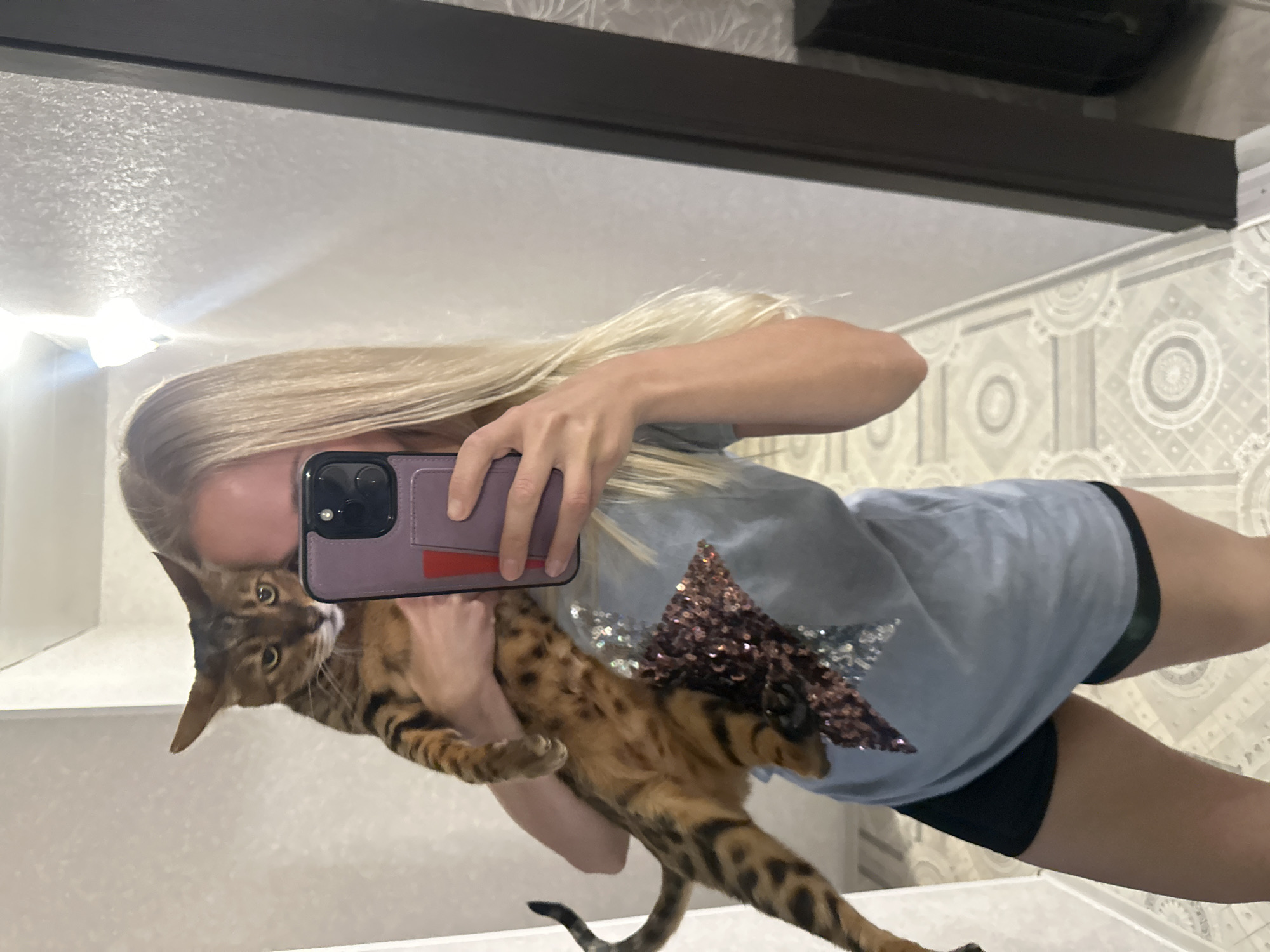 A woman with blonde hair is standing in a hallway, taking a mirror selfie while holding a Bengal cat. The woman is wearing a light blue t-shirt with a sparkly star design and black shorts. The cat is brown with black spots and stripes. The hallway has patterned wallpaper and tiled flooring. A dark brown door frame is visible on the right side of the image. The location is identified as Energetik, Russia.