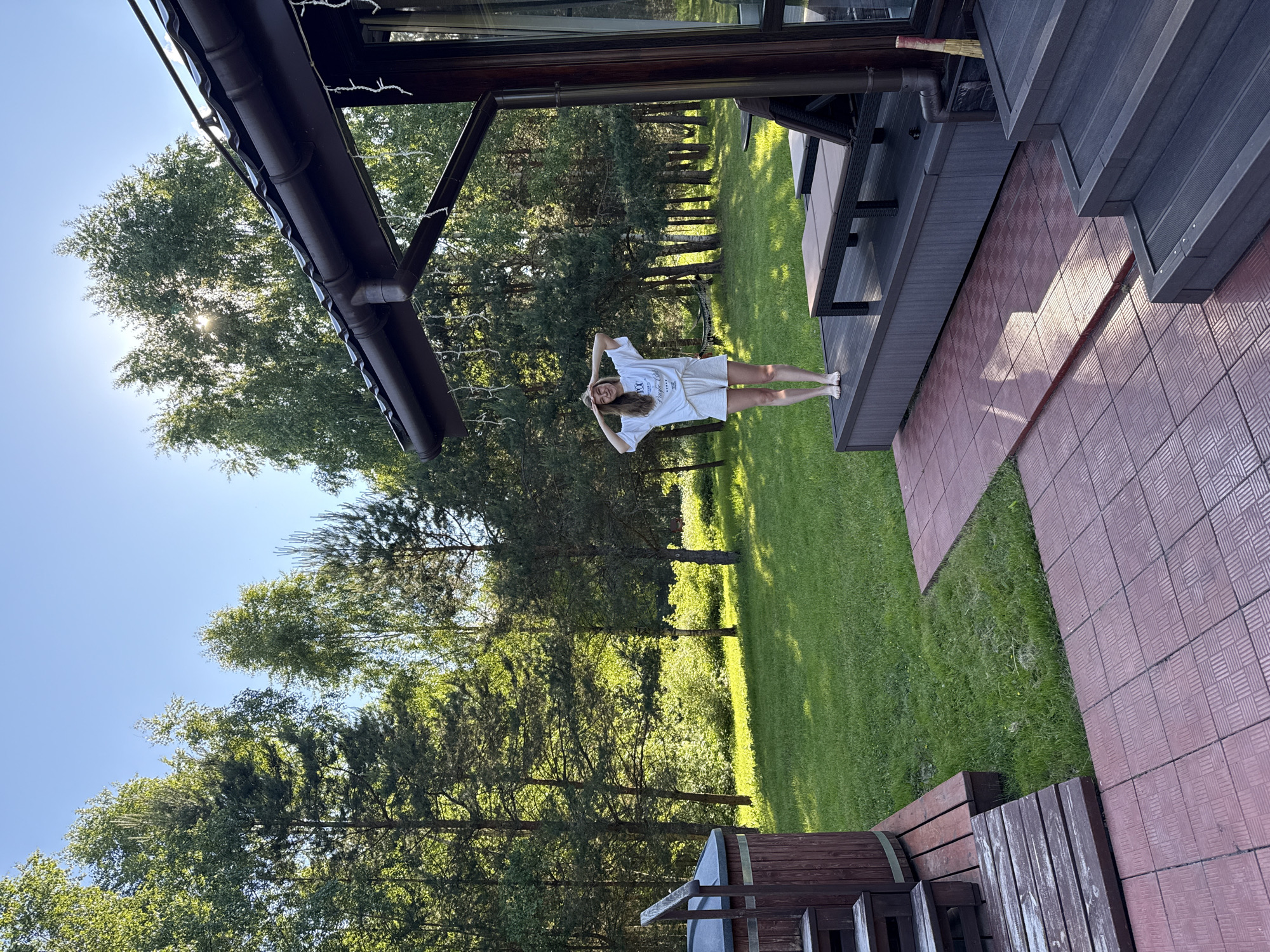 The image captures a bright, sunny outdoor scene, likely at a private residence in Копнино, Russia. A young woman with light brown hair stands barefoot on a dark wooden patio or deck. She is wearing an oversized white t-shirt with partial text that appears to read "100% ORGANIC ESSENTIALS" and light-colored shorts. Her arms are raised and bent, hands near her head, in a relaxed pose, and she is smiling.
To her right, parts of a modern house or outdoor structure made of dark wood or composite material are visible, featuring a gutter system. A red-brown patterned tiled pathway extends from the patio. The ground beyond the patio and pathway is a vibrant green lawn that stretches towards a dense backdrop of tall trees, including both coniferous and deciduous species, indicating a wooded area. Bright sunlight filters through the tree canopy, casting dappled light and shadows on the grass. The sky above is clear and blue. The overall atmosphere suggests a peaceful, warm day in a natural setting.