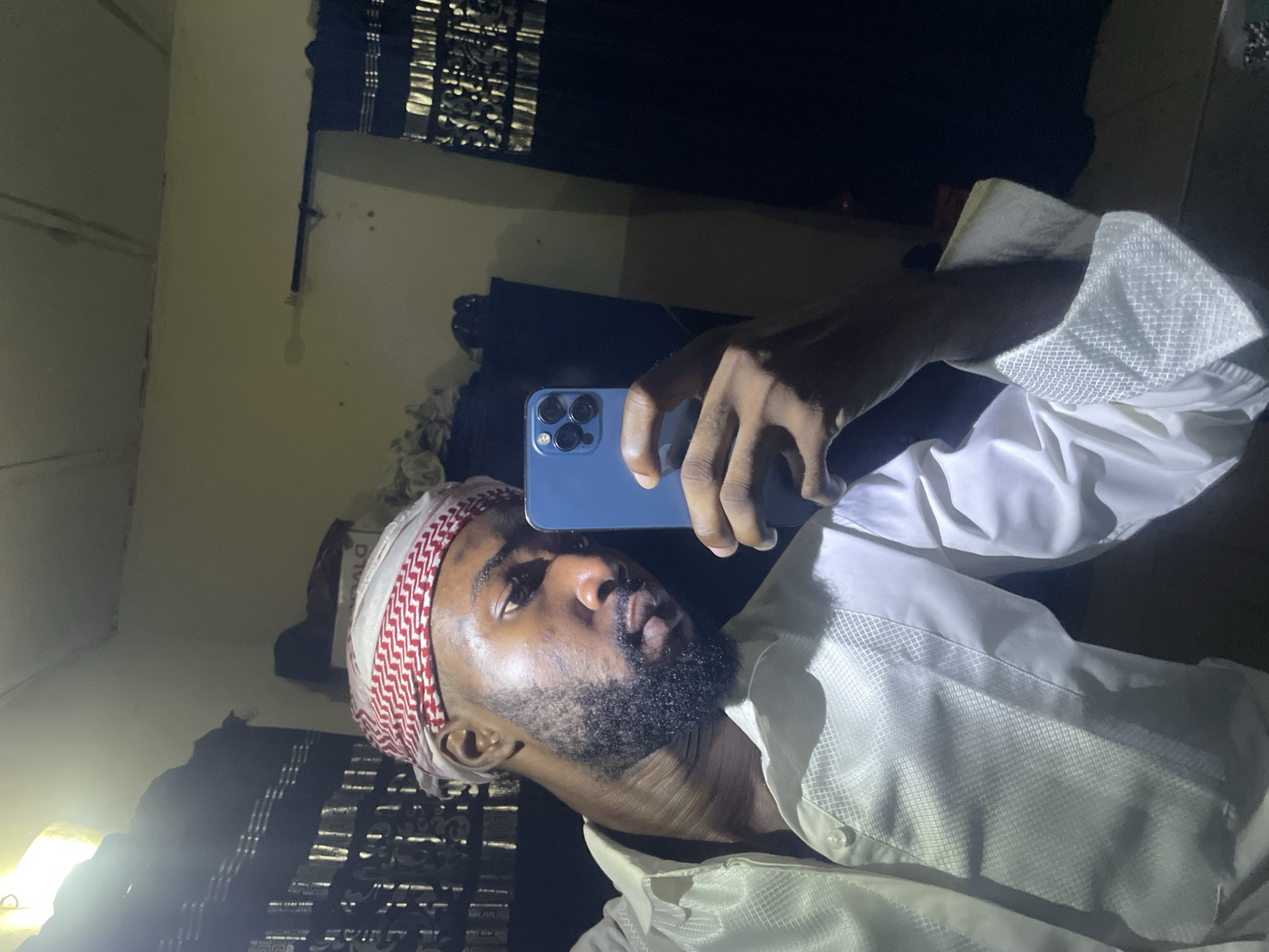 A close-up, low-angle shot depicts a man taking a selfie with a blue smartphone. He is wearing a white shirt with a patterned collar, and a red and white patterned head covering. He has a dark beard. The background is dark, but glimpses of a window covering and possibly other furniture can be seen. The location is Unknown, Unknown.