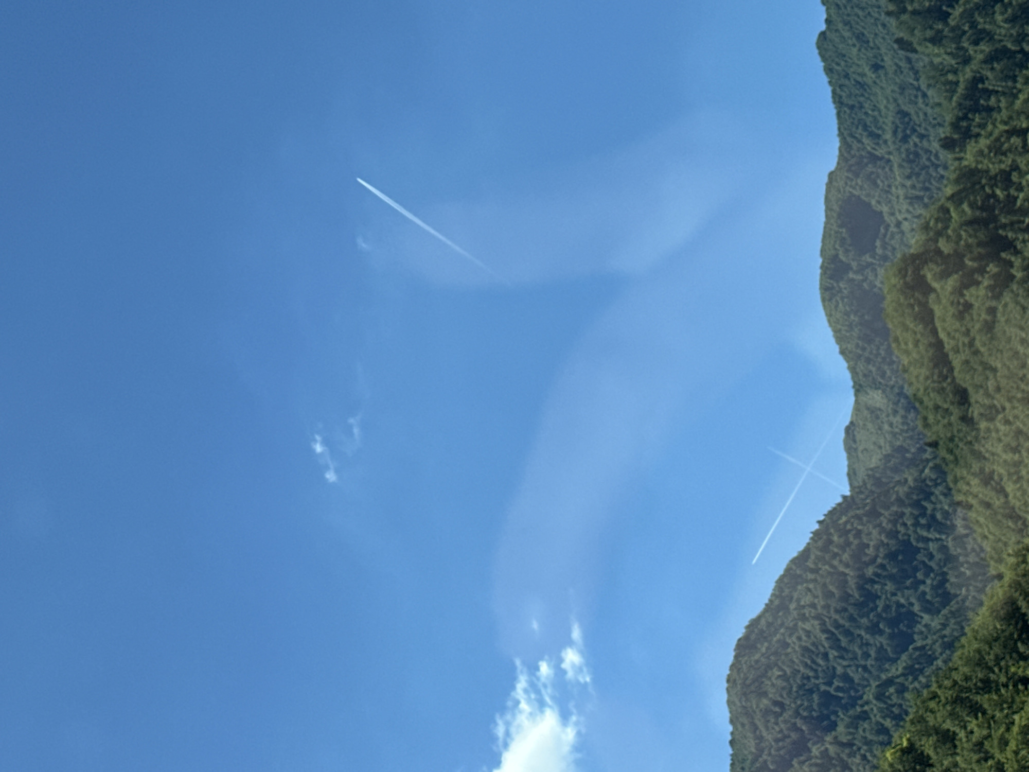The image shows a bright blue sky with two faint contrails from airplanes crossing each other. Below the sky are lush, green, tree-covered mountains, characteristic of the landscape in Klopot, Montenegro. The mountains form a textured, undulating line across the lower half of the image. There are no people or specific objects visible in the scene, only the natural environment of mountains and sky, with the transient marks of air travel.