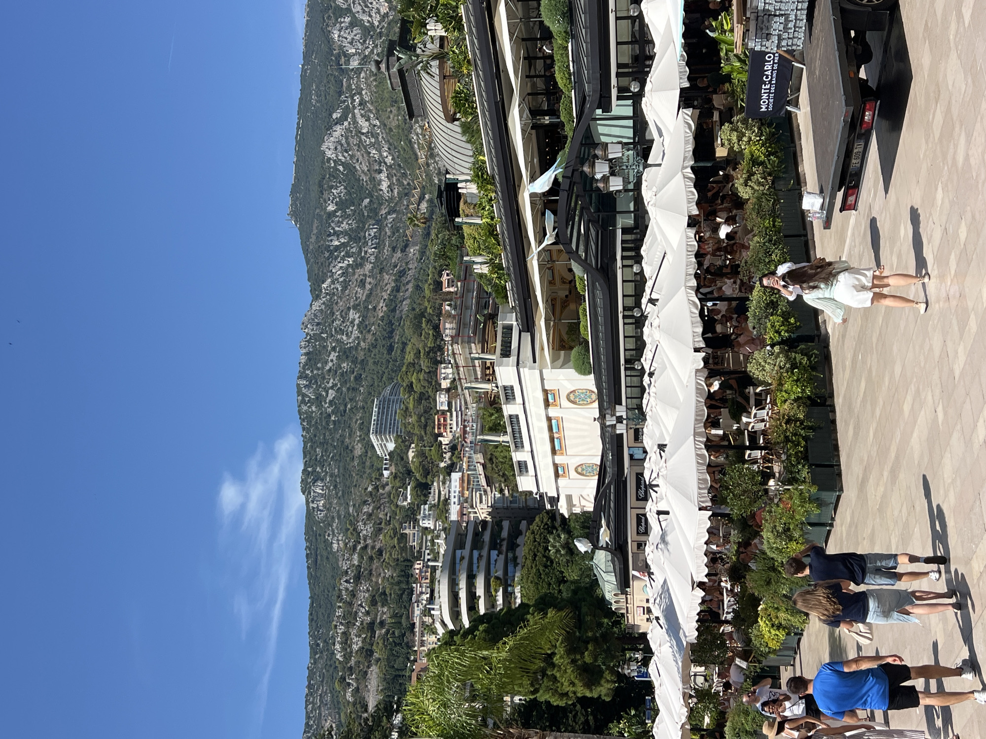 This image captures a sunny day in Monte Carlo, Monaco. The foreground features a paved walkway with several people strolling. To the left, a man in a blue t-shirt and black shorts walks away from the camera. Beside him, a young woman in a light blue denim skirt and dark t-shirt, and a young boy in dark shorts and a t-shirt, also walk away. Further into the frame, an outdoor dining area is set up with numerous white umbrellas covering tables where patrons are seated. Lush green plants and hedges surround the dining area, creating a vibrant, verdant atmosphere.
In the background, the iconic architecture of Monte Carlo unfolds. A large, ornate building with curved windows and decorative elements stands prominently. Behind this, the hilly landscape of Monaco rises, dotted with residential buildings, hotels, and verdant trees. The buildings are built on terraces, ascending the hillside. The sky above is a clear, bright blue, with a few wispy clouds. A flatbed truck with the text "MONTE-CARLO SOCIÉTÉ DES BAINS DE MER" is parked on the right side of the frame. Two bird sculptures, one light blue and one white, are perched on an awning above the dining area. The overall scene suggests a lively, upscale urban environment in the principality.