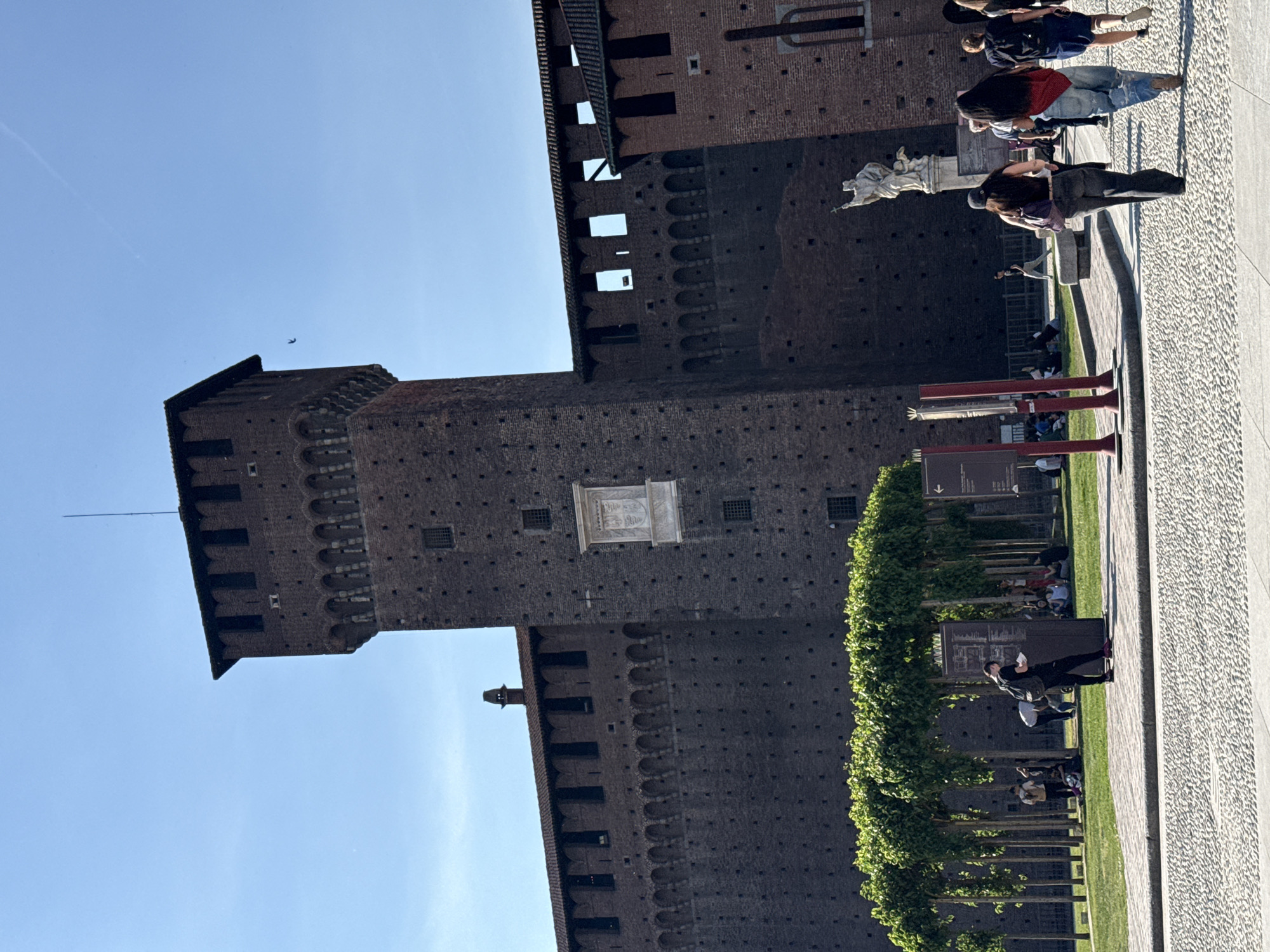 The image, rotated 90 degrees clockwise, captures a broad view of a large, dark brick castle under a clear blue sky, characteristic of Sforza Castle in Milan, Italy. The formidable structure dominates the left side of the frame, showcasing tall towers, crenellated battlements, and various window openings. A distinctive white stone relief sculpture is embedded within one of its walls, and a slender flagpole or antenna extends from the leftmost tower.
On the right, a wide, light-colored paved area, possibly cobblestones, stretches alongside the castle. Numerous people are present, enjoying the sunny day. In the mid-ground, a person in dark attire stands near a row of neatly trimmed green trees, possibly examining a handheld item or a dark informational sign. Further back, near a classical white statue on a pedestal, a small group of people can be seen, including an individual with long dark hair wearing a red top and blue jeans. Other visitors are visible sitting on grassy areas or benches beneath trees in the background.
Two red, T-shaped display structures are positioned in the mid-ground, suggesting an outdoor exhibition or information display. The bright natural light indicates it is daytime with clear weather. People are engaged in leisurely activities such as walking, standing, and relaxing. While signs are present, no text is clearly legible.