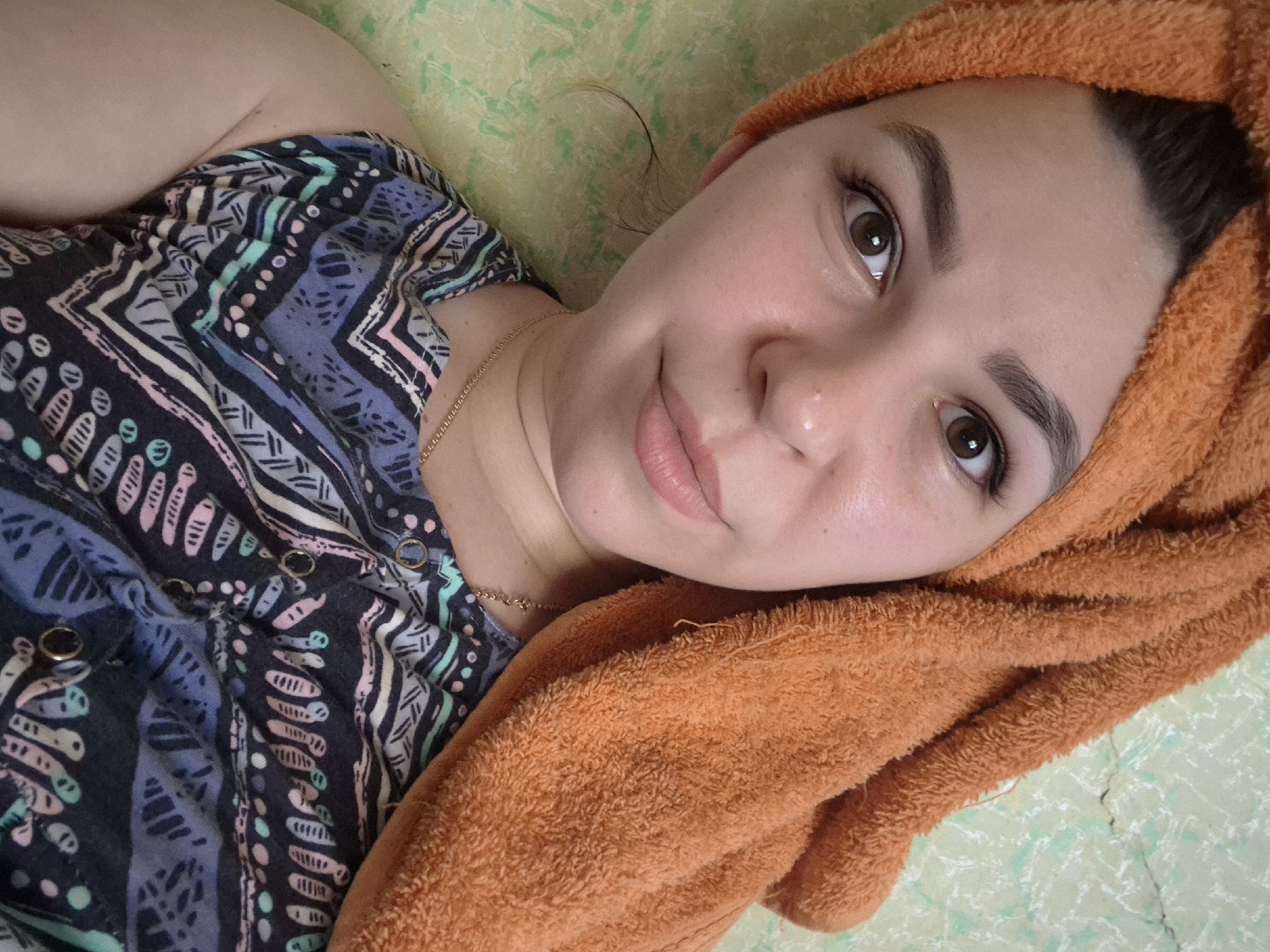 A young woman with brown eyes and dark eyebrows is shown from the chest up. She is wearing an orange towel wrapped around her head and a patterned sleeveless top. She is looking directly at the camera with a slight smile. The background appears to be a textured wall. There is no visible indication of the city of Kharnavy, Russia.