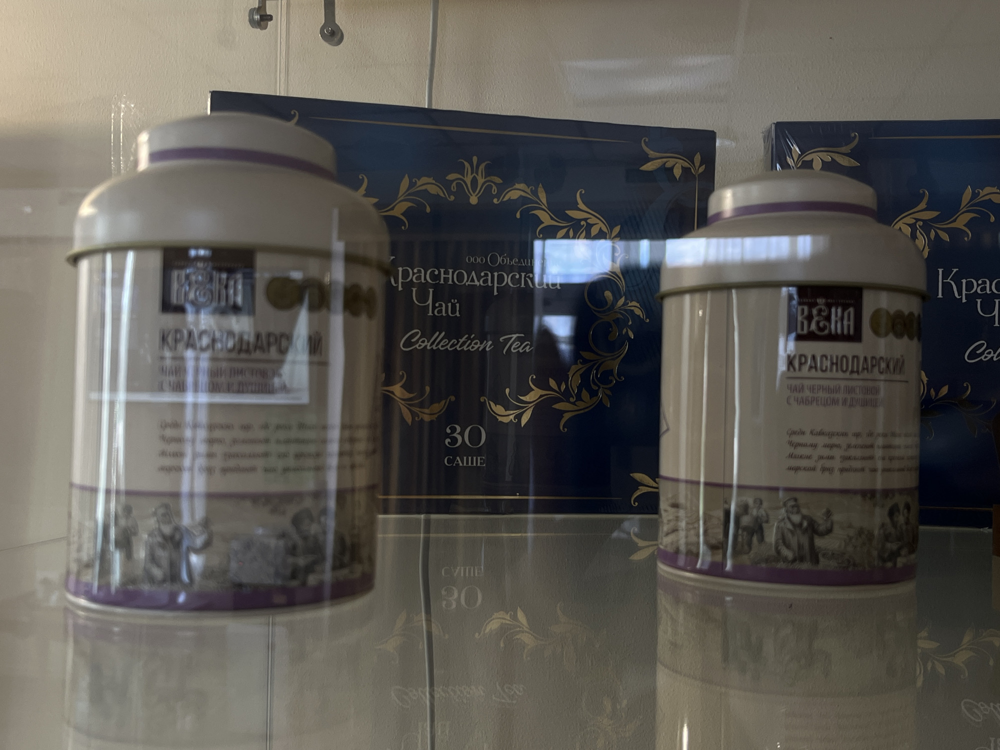This is a close-up, eye-level, indoor shot of a display case containing several tins of tea and their accompanying packaging. The background is a neutral, light-colored wall. Two dark blue boxes with ornate gold scrollwork and text are visible. The text on the boxes reads "ООО Объединение" and "Краснодарский Чай Collection Tea", with a number "30" and the word "САШЕ" (sachet) below it. Two cylindrical tea tins, one in the foreground on the left and another slightly behind and to the right, are also displayed. The tins are a light cream color with a purple band around the top. They feature labels with similar gold scrollwork and text. The labels on the tins include "КРАСНОДАРСКИЙ" and "ЧАЙ ЧЕРНЫЙ ЛИСТОВОЙ С ЧАБРЕЦОМ И ДУШИЦЕЙ". Below this text, there are intricate illustrations of people in what appears to be a historical or rural setting. The tins are reflecting the surrounding environment, including parts of the boxes and the light source. The display case has a reflective surface, as evidenced by the reflection of the tea tins and their labels at the bottom of the image. The scene suggests a commercial display, likely within a shop or museum, exhibiting Krasnodar tea.