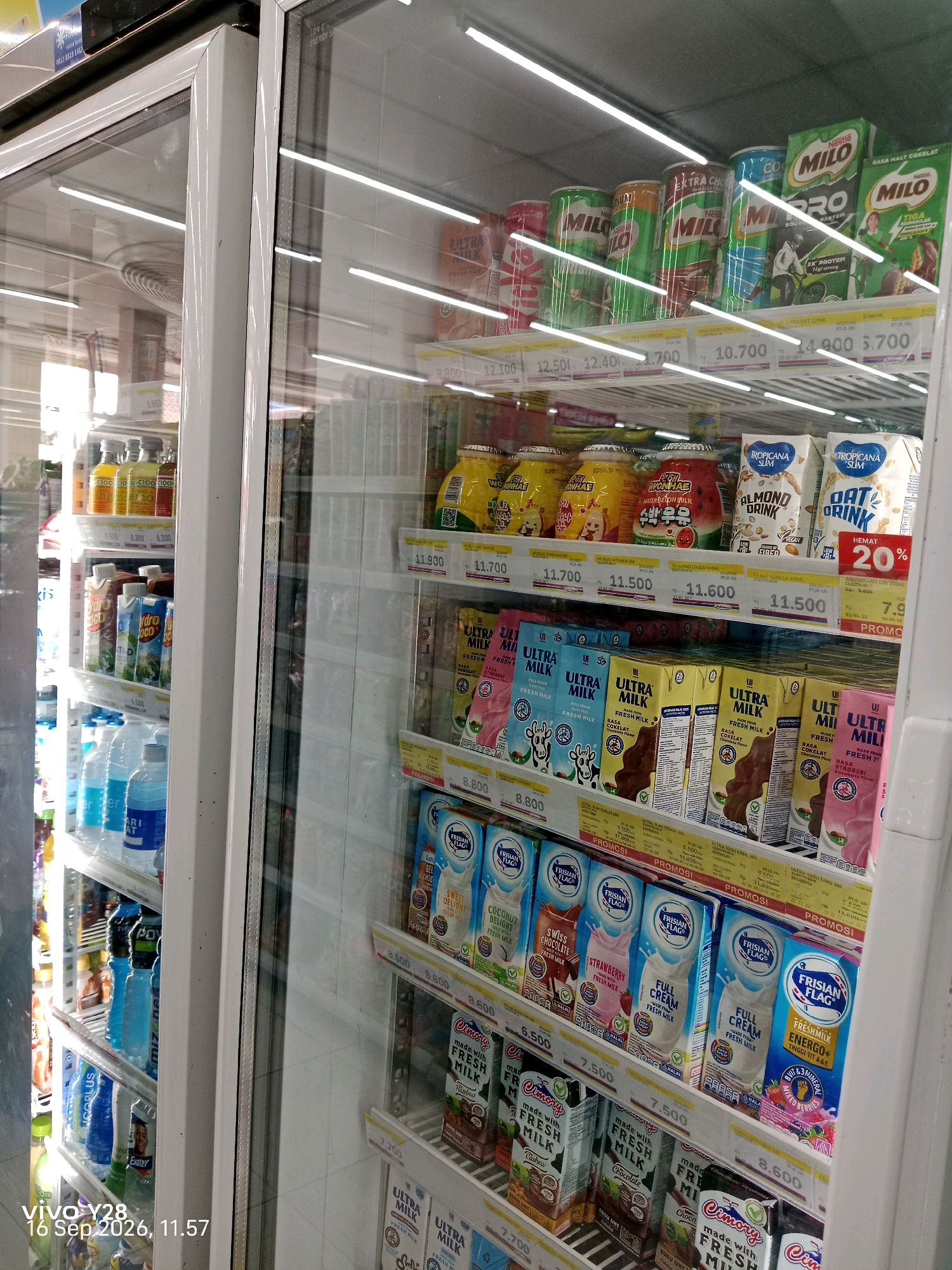 The image shows a refrigerator display case in a convenience store in Tlogosari, Indonesia. The shelves are stocked with various beverages, including milk cartons, bottles of water, and canned drinks. Products like Milo, Ultra Milk, Tropicana Slim, Frisian Flag, and Cimory are visible. Prices are displayed below some of the products. The lighting in the store is bright, and reflections are visible on the glass doors of the refrigerator. The overall environment appears clean and organized, typical of a retail setting. There are no people visible in the image. The timestamp "vivo Y28, 16 Sep 2026, 11:57" is present in the bottom left corner.