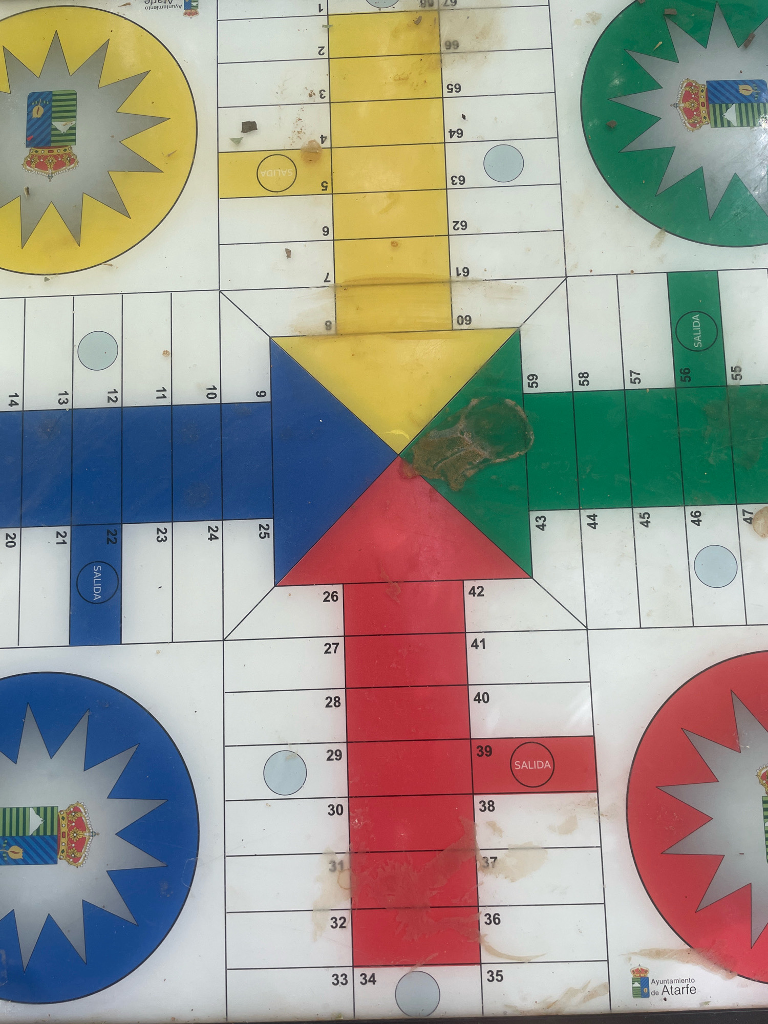 The image shows a close-up, top-down view of a Ludo board game, likely outdoors, given the presence of dirt and debris on the surface. The board is divided into colored sections with numbered squares, and features circular "home" areas with emblems. One of the emblems visible is from the "Ayuntamiento de Atarfe," indicating the game is located in Atarfe, Spain. There are no people or animals visible in the image. The board appears worn and used, with visible smudges and dirt in various sections. The game is set up on a flat surface, with the overall scene being a static, overhead shot of the game board.