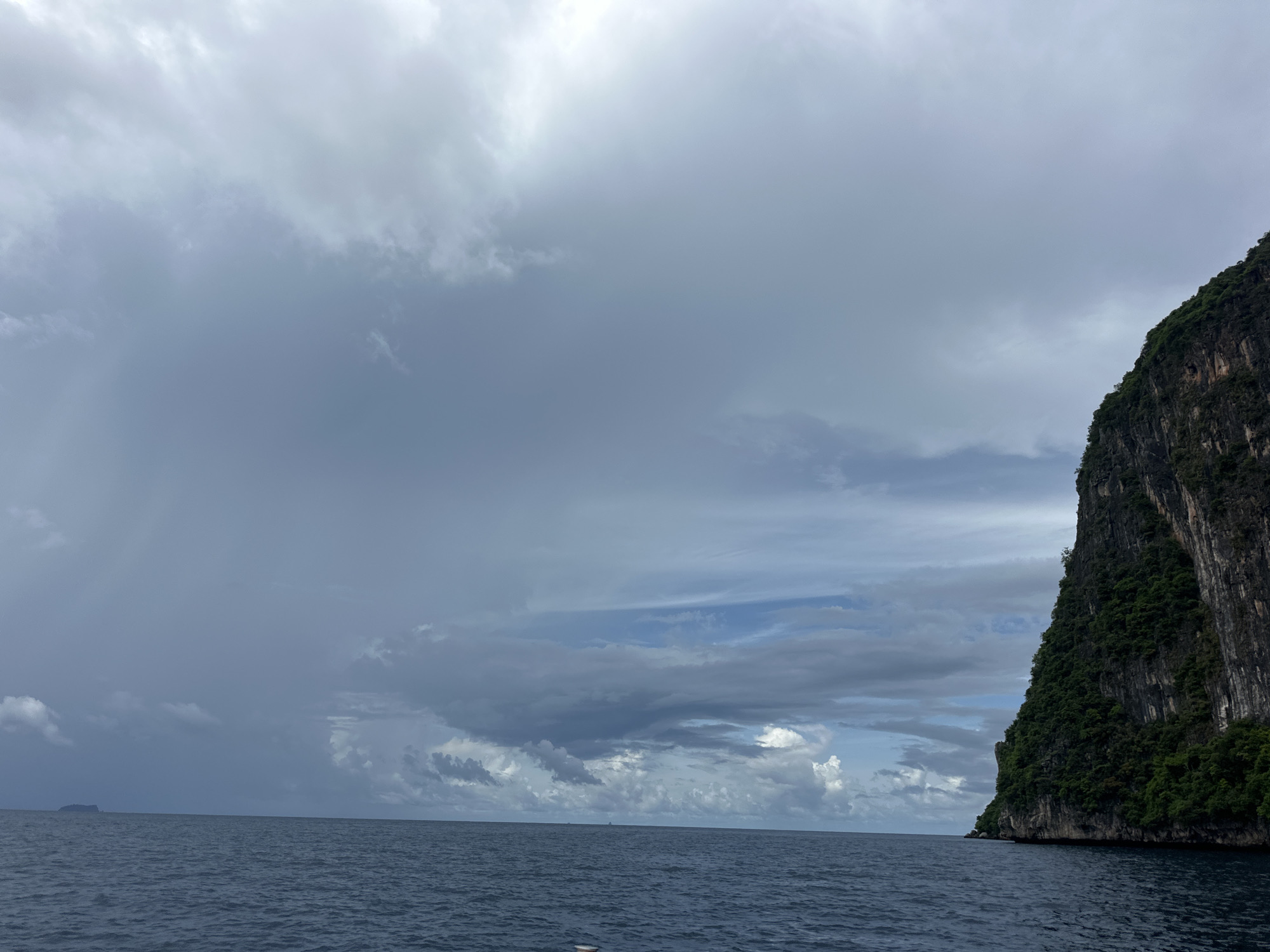 The image depicts a seascape with a large, sheer cliff face on the right side, covered in dense green vegetation. The cliff rises dramatically from the dark blue ocean. The sky above is heavily overcast with a mixture of dark gray storm clouds and lighter patches of blue sky peeking through. In the distance, a small, dark island is visible on the horizon to the left. The water appears choppy. This scene is likely located in the vicinity of Haad Hin Khom, Thailand, a region known for its dramatic karst landscapes and ocean views. There are no people or specific actions depicted in the image; it is a natural landscape view.