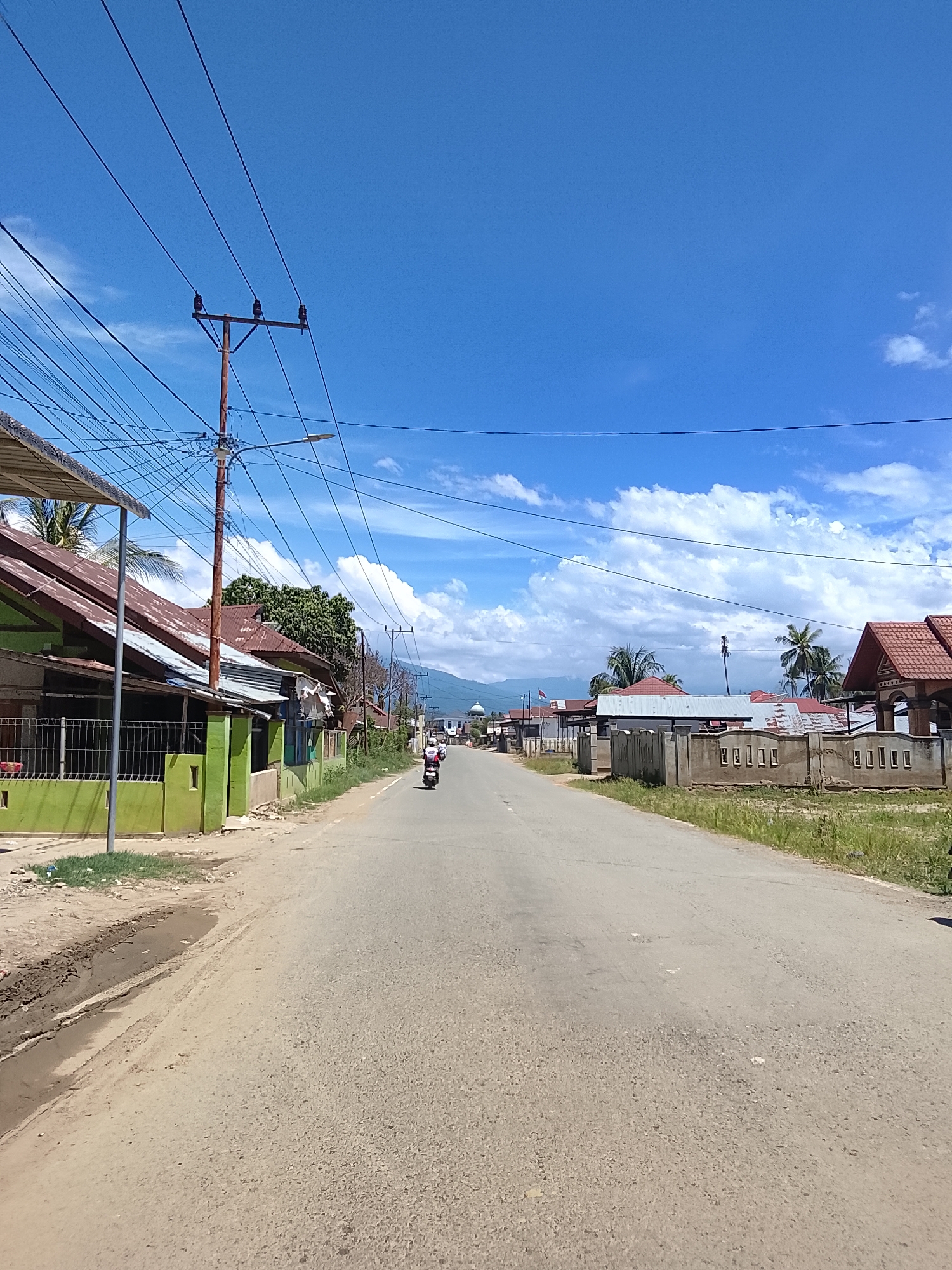 This image depicts a street scene in Blang Cut, Indonesia, under a bright, partly cloudy sky. The asphalt road stretches into the distance, with a dirt shoulder on the left and grassy verges on the right. Several houses with corrugated metal roofs line the left side of the road, one of which is painted bright green. Power lines crisscross the sky, supported by wooden utility poles, one of which has a street lamp attached. Two individuals on a motorcycle are traveling away from the camera down the road. In the distance, mountains are visible, and a dome-shaped structure, possibly a mosque, can be seen on the right side of the road. The overall impression is a sunny, typical day in a semi-rural or suburban Indonesian town.