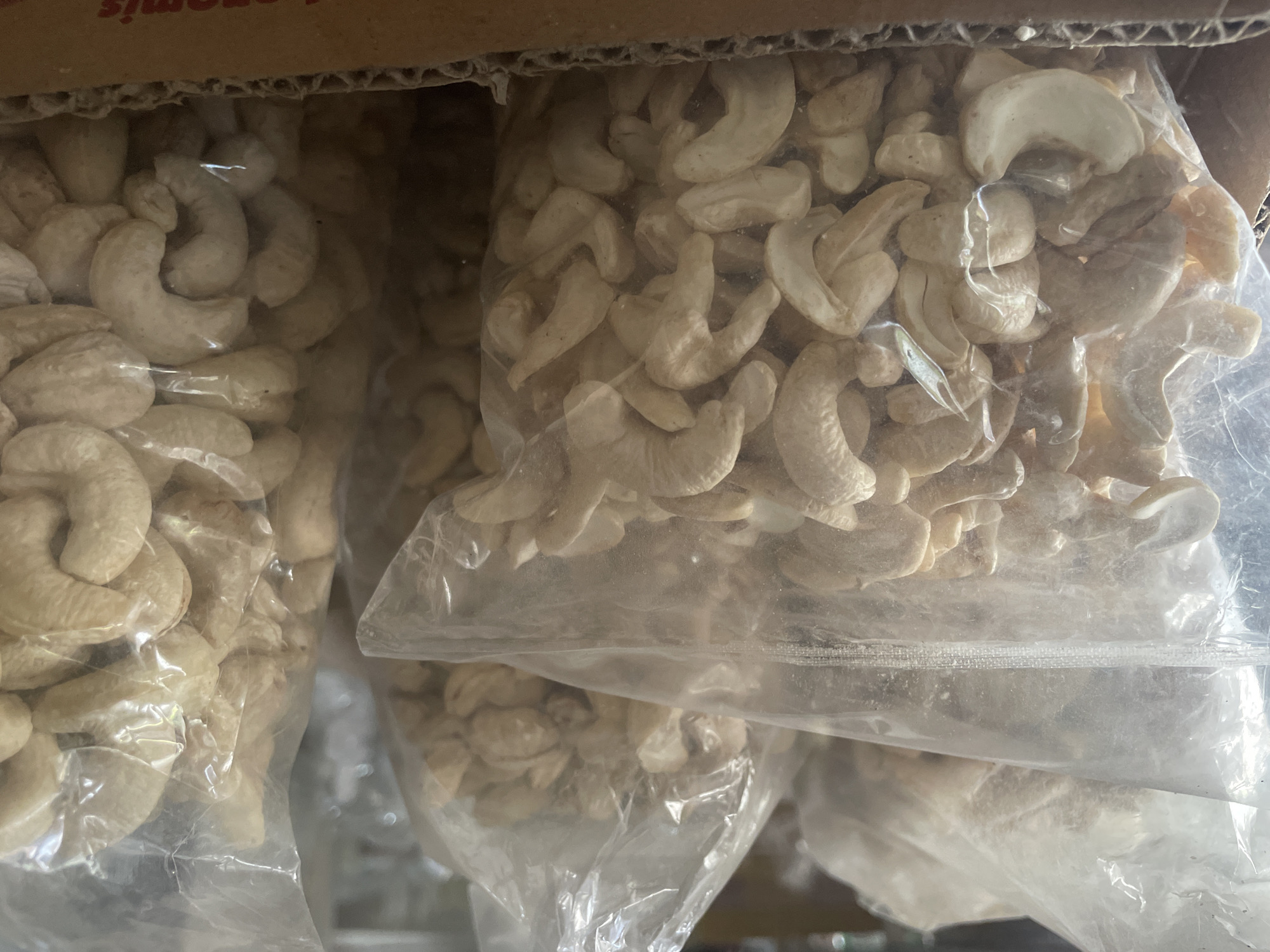 The image shows several plastic bags filled with raw cashews. The bags are stacked in a cardboard box, and the scene appears to be a retail or storage environment. The cashews are light beige in color and have the characteristic kidney shape. The plastic bags are transparent, allowing the cashews to be clearly visible. The lighting in the image is somewhat dim, casting shadows on some of the cashews. There is a partial view of a cardboard box in the foreground, with some text visible on it. The background is blurred but suggests a shelving unit or more products. The image does not provide any specific information about the city of Purworejo, Indonesia, or any people present.