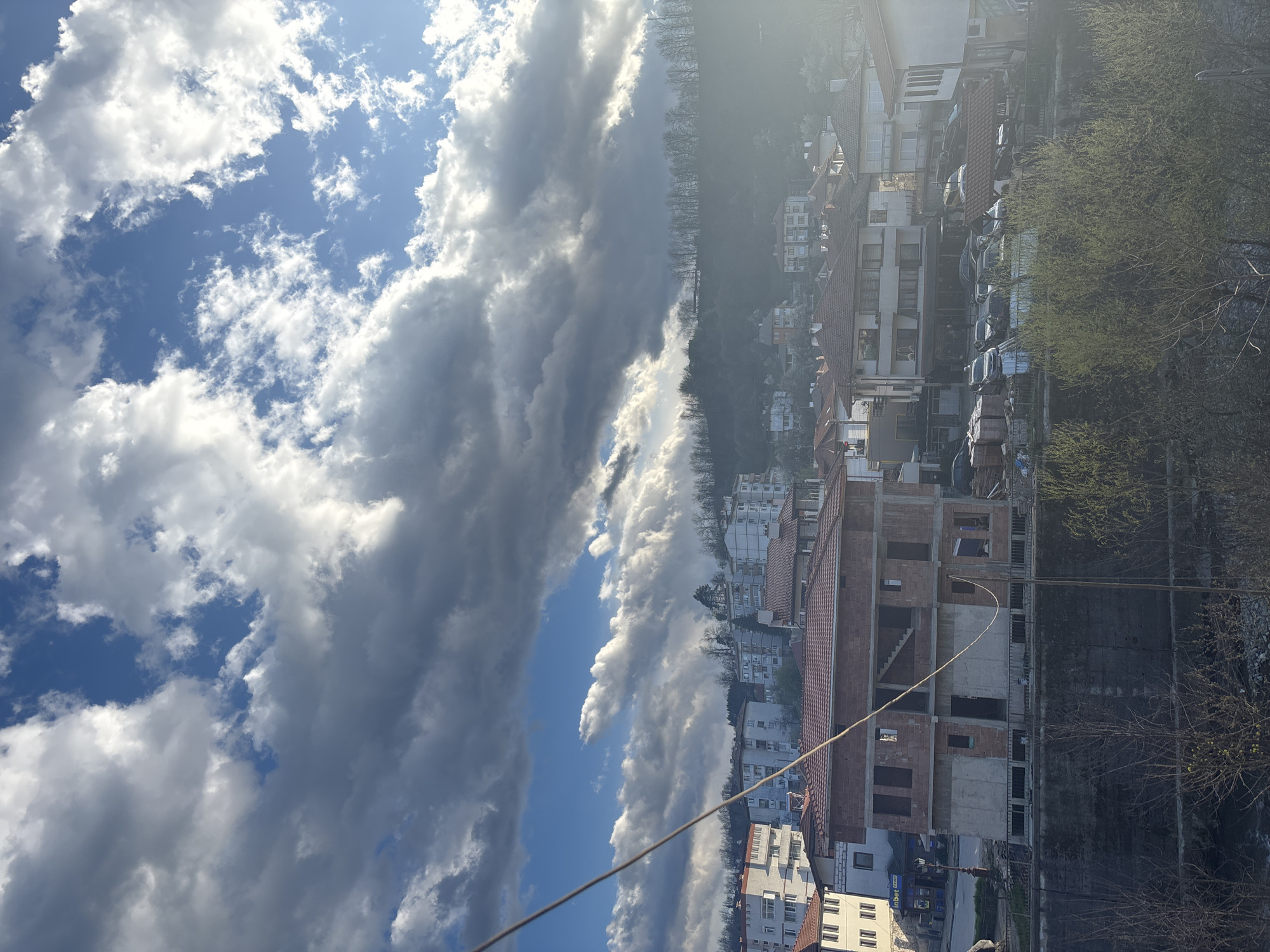The image shows an upward-angled view of a cityscape, likely taken from a high vantage point in Zlatograd, Bulgaria. The sky dominates the left portion of the frame, exhibiting a mix of blue sky and dense, white and grey clouds. A building under construction, characterized by its red brick and concrete structure, is prominently featured in the right foreground. Various residential and commercial buildings with red tile roofs are visible in the background. Trees with budding green foliage are interspersed throughout the scene. A power line diagonally cuts across the lower portion of the image. Overall, the scene conveys a blend of urban development and natural elements in a Bulgarian city setting.