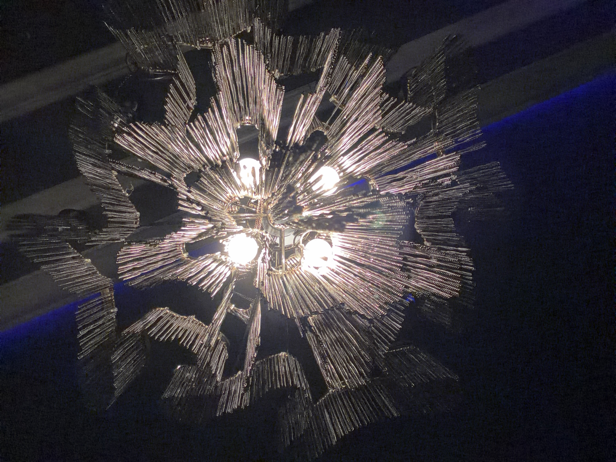 A chandelier composed of a metal frame with numerous metal rods radiating outwards is illuminated by several light bulbs in the center. The background is dark, with a blue light source visible near the top right. The location is unknown.