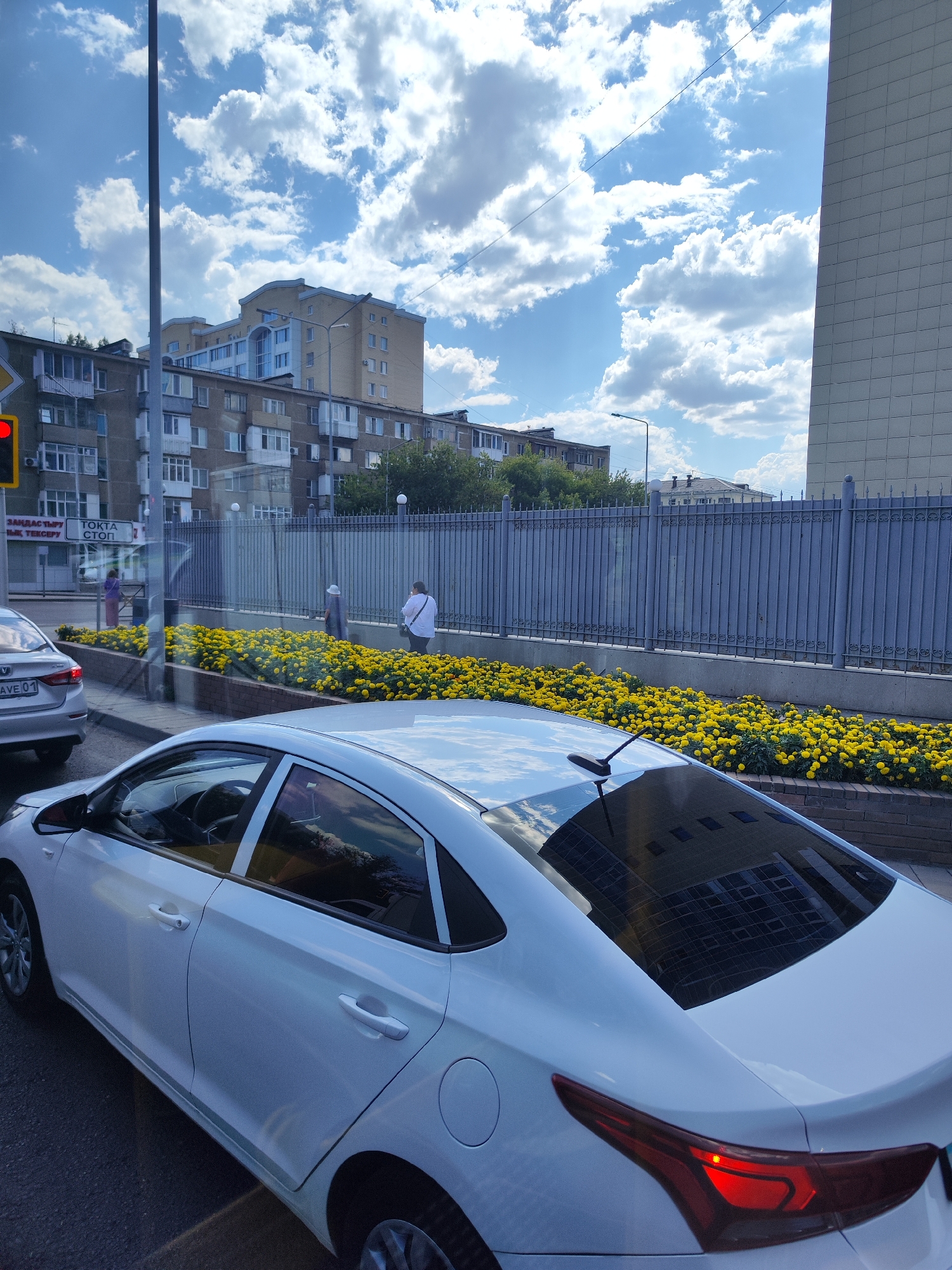 In Astana, Kazakhstan, on a bright, cloudy day, a white sedan is parked on the street, its rear taillight illuminated red, suggesting it is either braking or has its hazard lights on. The car is positioned on the right side of the frame, with its rear and right side visible. In the background, across a fence, a flower bed filled with vibrant yellow flowers is bordered by a brick wall. Beyond the flowers, several people are visible, some walking and one standing near a building with a blue fence. In the distance, apartment buildings and a modern structure with a tiled facade dominate the skyline under a dynamic sky with scattered white clouds. A traffic light with a red signal is also visible on the left side of the image, indicating a potential traffic stop. The scene appears to be captured from another vehicle, as evidenced by reflections on the car windows.