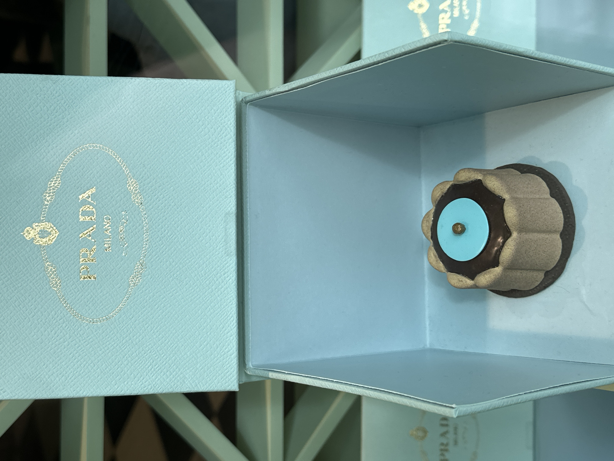 A close-up shot reveals a light blue Prada box, partially opened, containing a decorative object inside. The box lid, positioned on the left, showcases the Prada logo embossed in gold. Inside the box, affixed to the wall, is a circular ornament resembling a flower, with a tan petal-like outer ring, a brown central section, and a turquoise blue dot in the middle. The box itself appears to be part of a display, possibly in a retail setting, with glimpses of other similar boxes and a light-colored grid-like structure in the background. The location is suggested to be in London, United Kingdom.