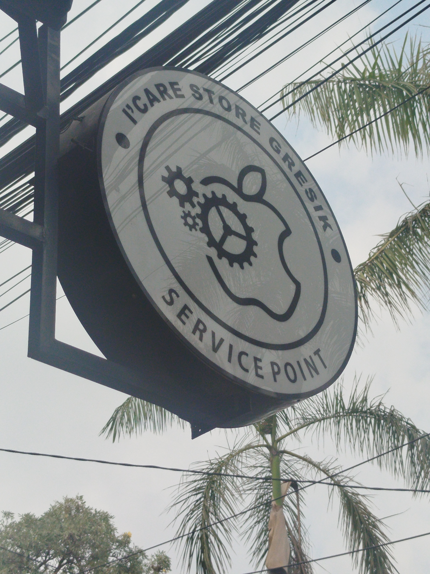 The image shows a circular sign for "I'CARE STORE GRESIK SERVICE POINT" hanging from a metal support structure. The sign features a stylized Apple logo with gears inside. Palm trees and overhead wires are visible in the background, suggesting an outdoor urban or semi-urban environment. The sky is overcast. The sign is positioned at an angle, facing slightly upwards. The location is specified as Yosowilangun, Indonesia.