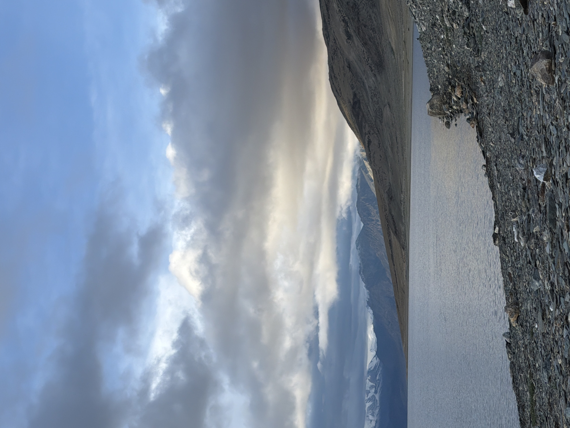 An outdoor, low-angle, vertical shot captures a serene landscape of a body of water with a rocky shore and a mountainous backdrop. The water, a muted blue-gray, reflects the overcast sky. The shore is densely covered with small, dark gray stones and larger rocks, some of which are lighter gray or white. In the distance, rolling hills transition into rugged mountains, with snow-capped peaks visible on the far left. The sky above is a mix of blue patches and large, dramatic clouds in shades of gray and white, with sunlight breaking through in some areas, illuminating the clouds and the distant mountain slopes. The scene is devoid of human presence. The location is identified as a lake in Ladakh, India.