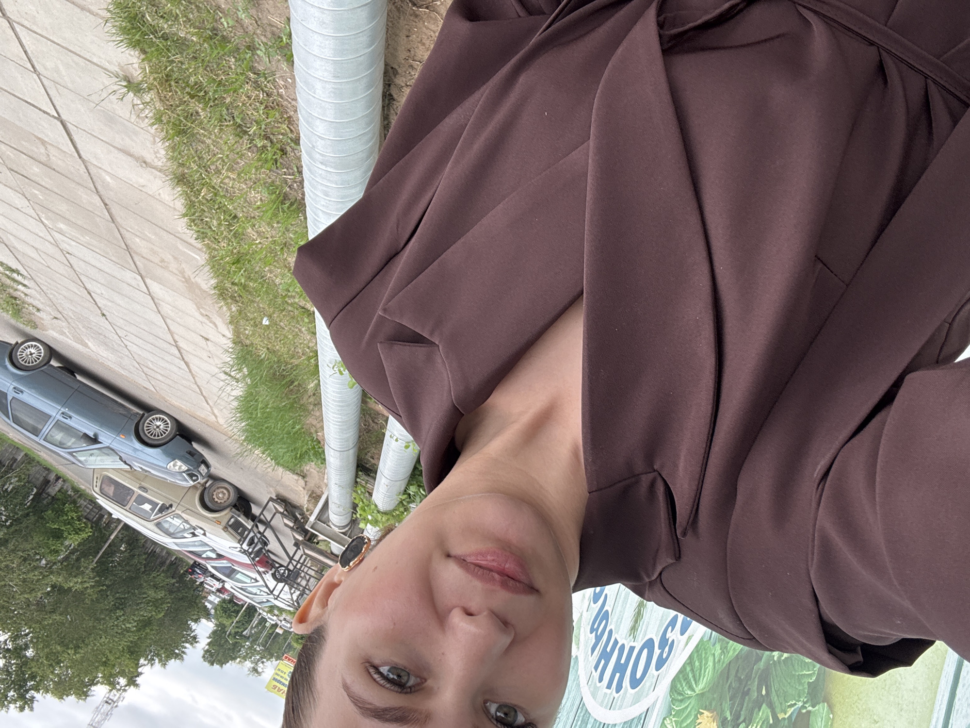 A woman takes a selfie outdoors in Chaikovsky, Russia. She is wearing a dark brown blazer with an asymmetrical neckline and a belt. She has on large oval earrings with a dark stone. In the background, cars are parked along a street, with trees and buildings visible further back. Two large pipes run along the ground in the foreground. The overall impression is that of a casual, candid moment captured on a street in a Russian city.