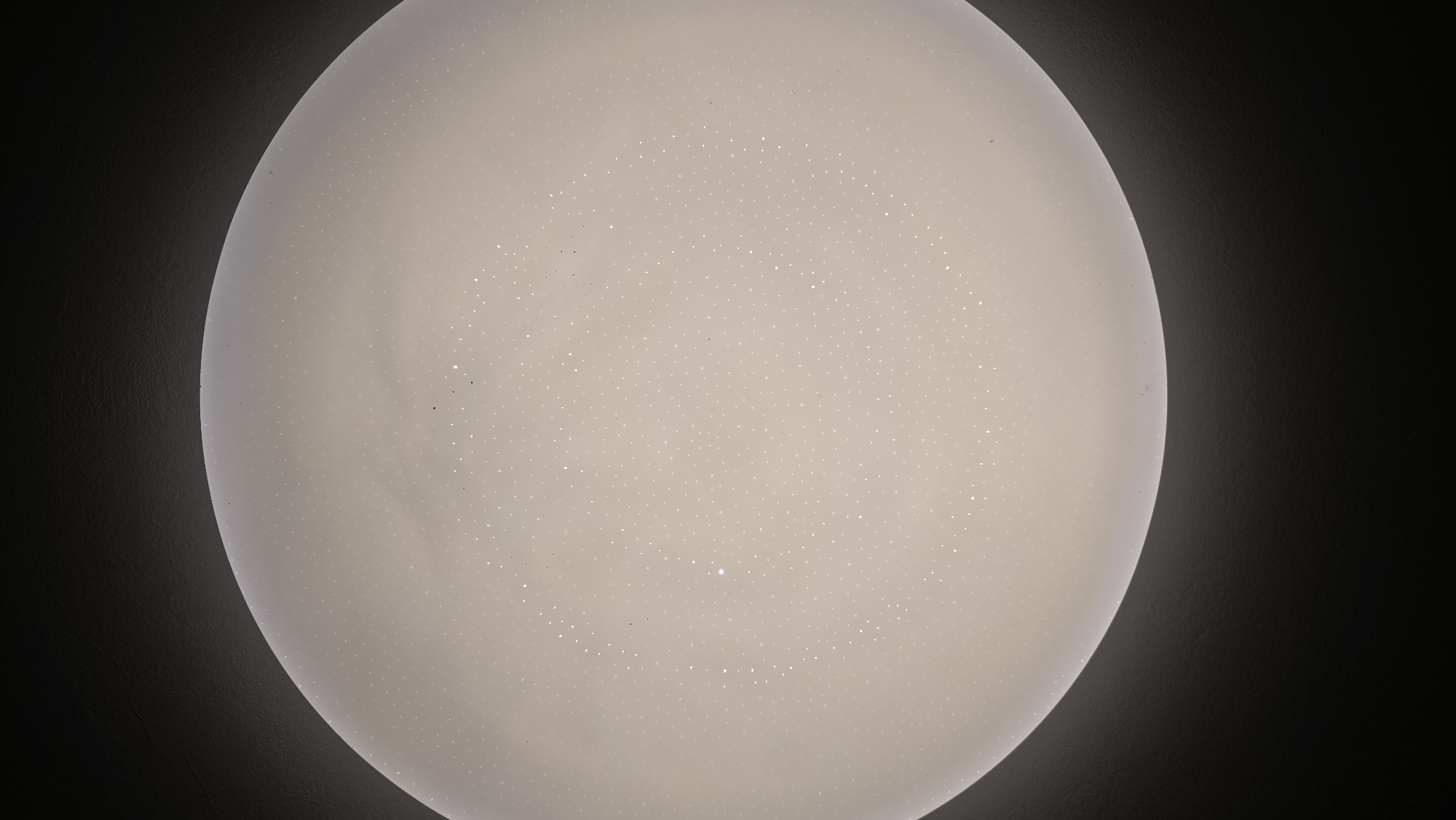 The image shows a close-up, downward-facing view of a white, circular ceiling light fixture. The fixture has a speckled or starry pattern of small, bright dots distributed evenly across its surface. The light is on, emitting a soft, diffused glow. The edges of the fixture are illuminated, and a dark, textured surface, likely a ceiling, is visible around the perimeter of the light. The overall impression is of a modern, decorative lighting element. There are no people or specific urban landmarks visible.