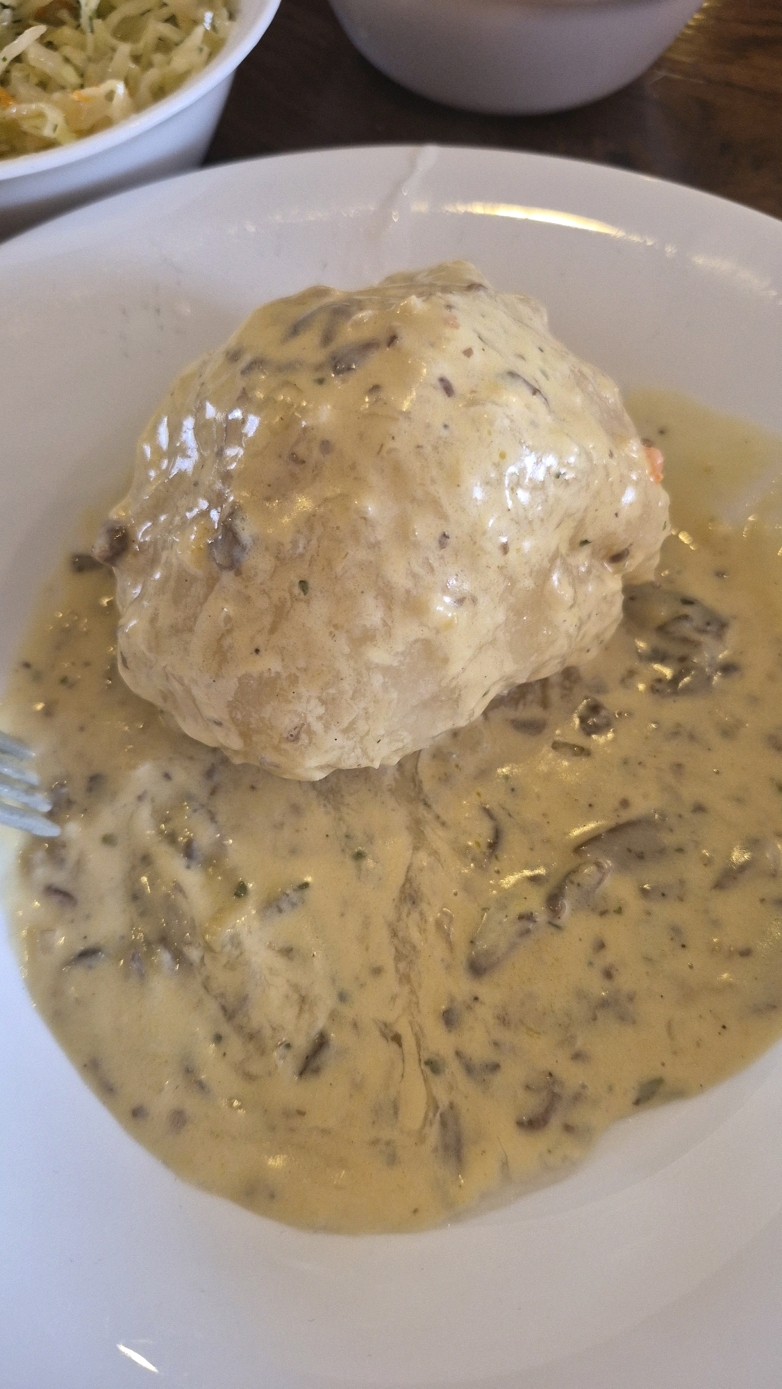 A close-up, overhead shot shows a dish of food, likely a meatball or dumpling, covered in a creamy, mushroom gravy. The food is served on a white plate, with a fork visible to the left of the plate. In the upper left corner of the image, a white bowl filled with coleslaw can be seen. The background is dark and out of focus. The food appears to be served in a restaurant setting in Smėlynė, Lithuania.
