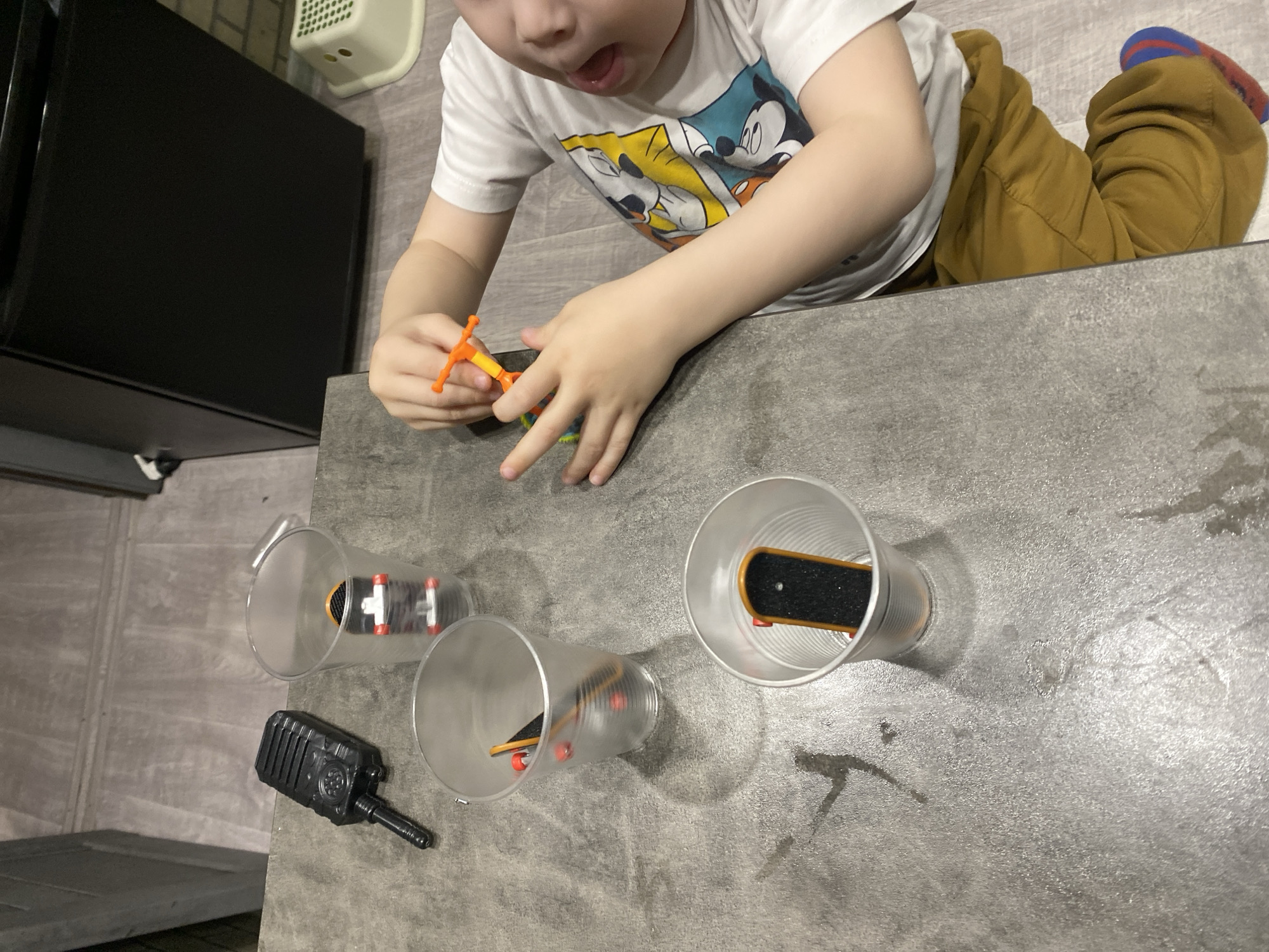 The image shows a child playing with toys on a gray surface. The child is wearing a white t-shirt with cartoon characters and tan pants. The toys include three plastic cups, each containing a finger skateboard. There is also a black toy walkie-talkie. The location is not identifiable as Sochi, Russia from the image.