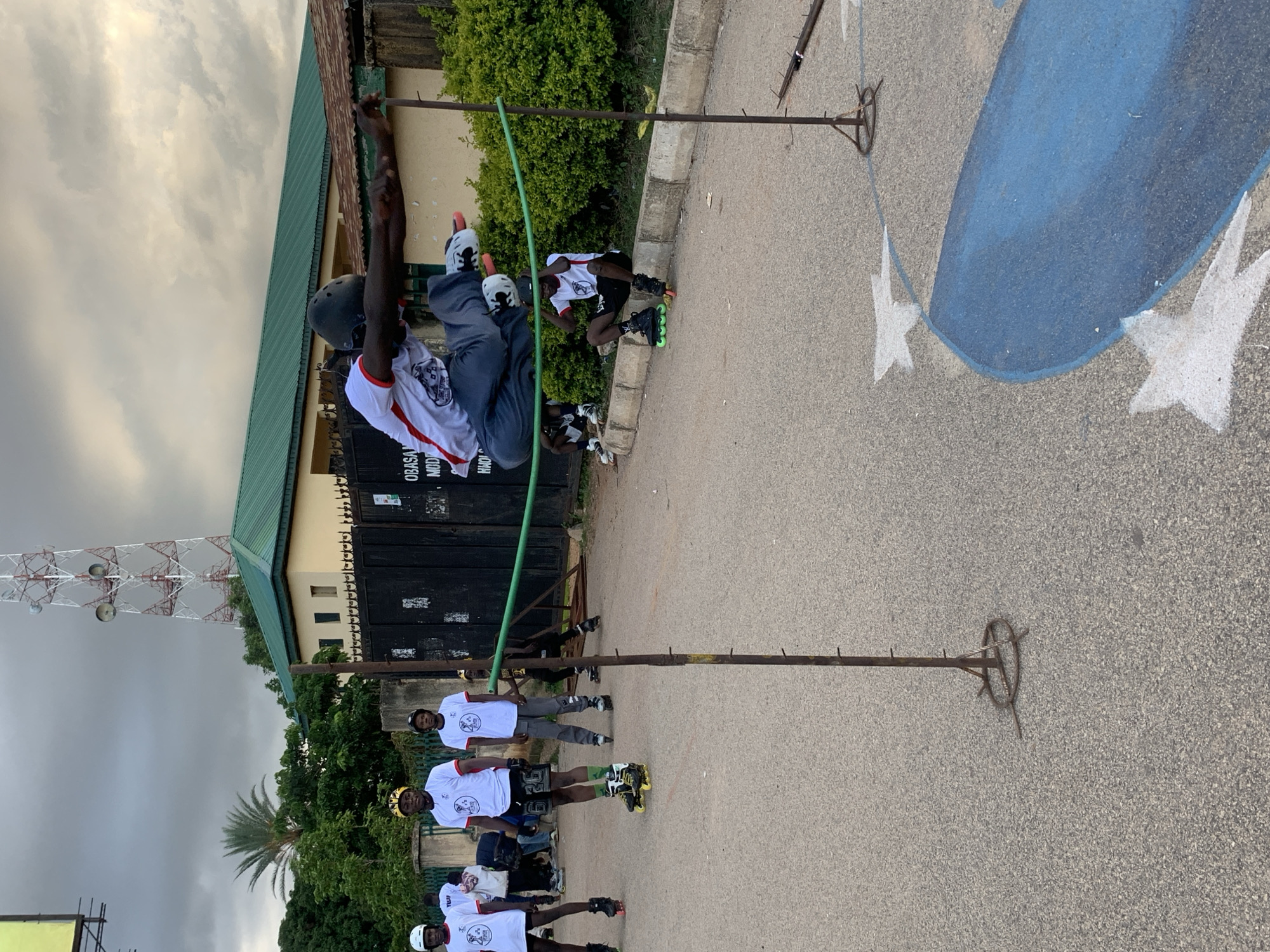 An overcast day in Jos, Nigeria, where a group of young men on rollerblades are practicing. The focal point is a young man in mid-air, performing a jump over a green rope suspended between two metal poles. He is wearing a white t-shirt with red trim, grey shorts, a black helmet, and rollerblades with black and white wheels. His body is arched, arms outstretched, and legs bent as he clears the obstacle.
To his left, three other young men in similar white t-shirts and helmets stand on rollerblades, observing. One is wearing yellow headgear, another black, and the third's headgear is not clearly visible. They are all wearing white t-shirts with a circular logo. The man on the far left is wearing black shorts, the middle man grey shorts, and the man on the right grey pants.
Further to the right, a young man is sitting on the curb in rollerblades, looking towards the jumper. He is wearing a white t-shirt and black shorts. Another person on rollerblades is partially visible in the background, near a dark metal gate.
The scene takes place on an asphalt road marked with white painted stars and a large blue shape, possibly part of a playground or sports area. Behind the participants, there is a green hedge and a building with a green roof. In the background, a prominent telecommunications tower with red and white markings stands against the cloudy sky, characteristic of the Jos landscape. The overall atmosphere suggests an outdoor training session or recreational activity.