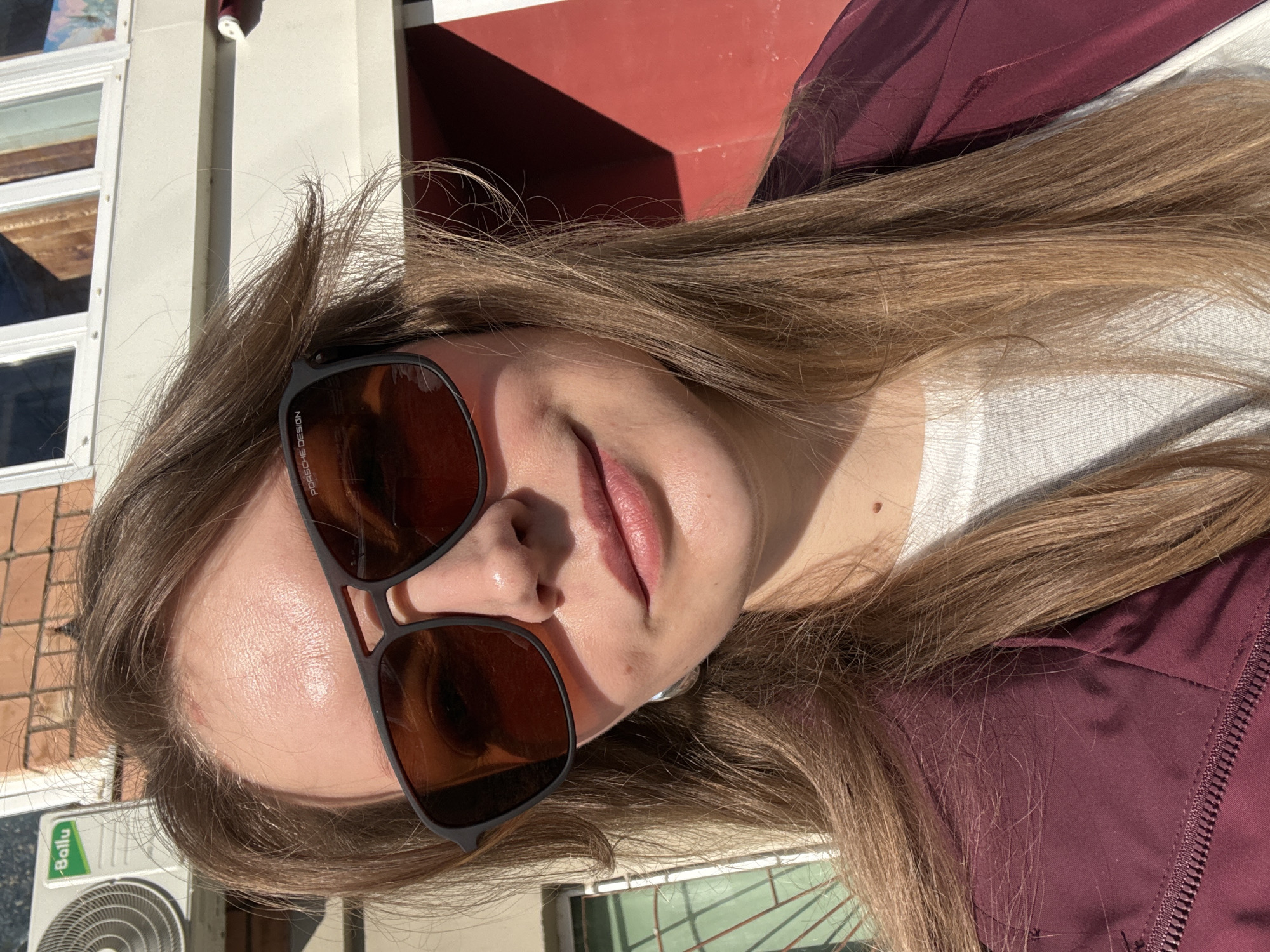 A young woman with long blonde hair poses outdoors, wearing sunglasses with "PORSCHE DESIGN" visible on the frame. She is wearing a white shirt and a burgundy jacket. The background includes a building with white trim and red sections. A "Bailu" branded air conditioning unit is visible in the lower left corner. The location appears to be in the city of Izhevsk, Russia.