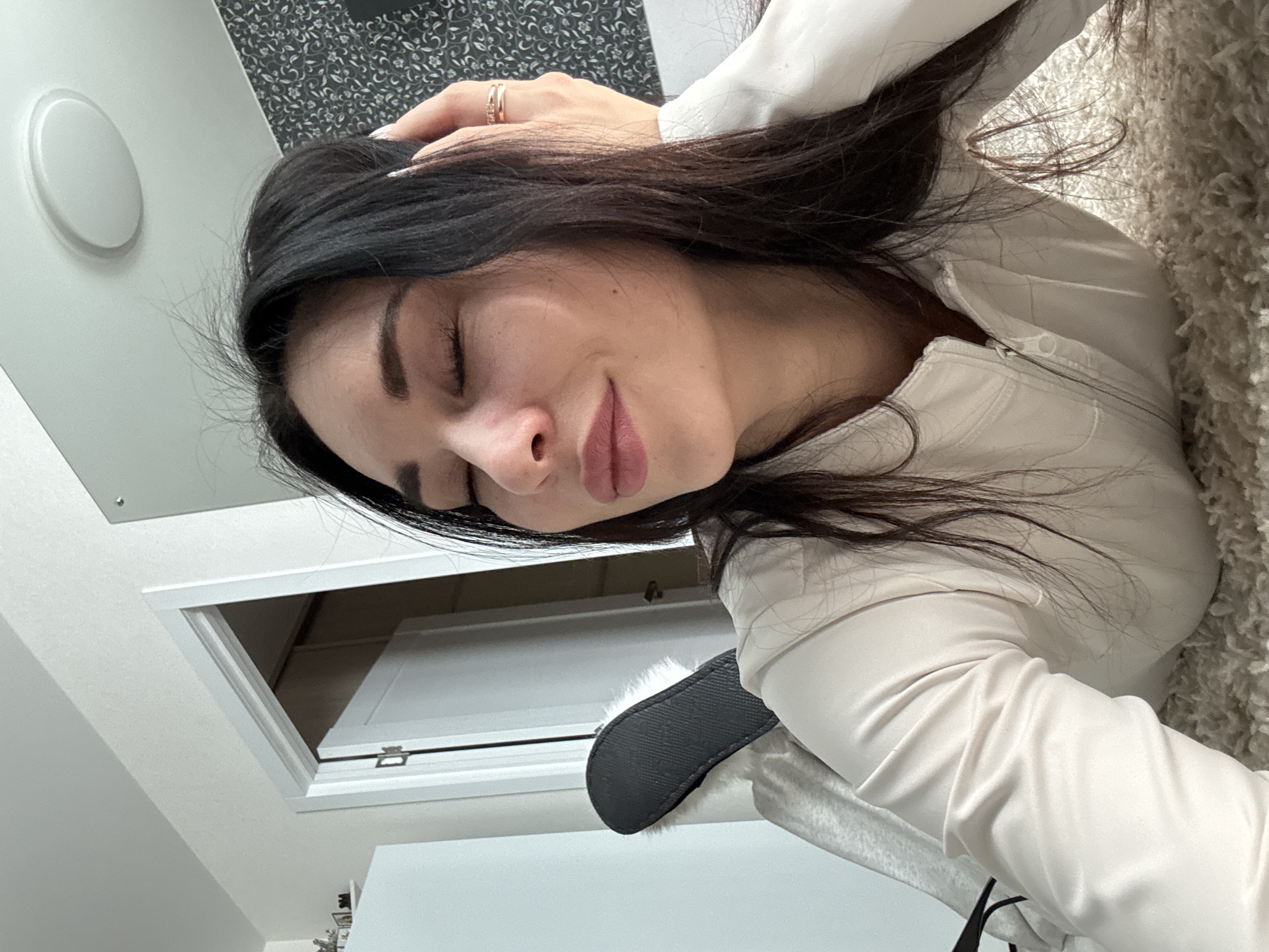 The image shows a woman with dark hair and closed eyes, wearing a white top. She appears to be resting her head on a light-colored, textured surface. A hand with a ring is resting on her head. In the background, there is a white wall with a circular light fixture and a dark patterned design. A partially open white door or hatch is also visible. There is no discernible information to conclude the location is Chaikovsky, Russia.