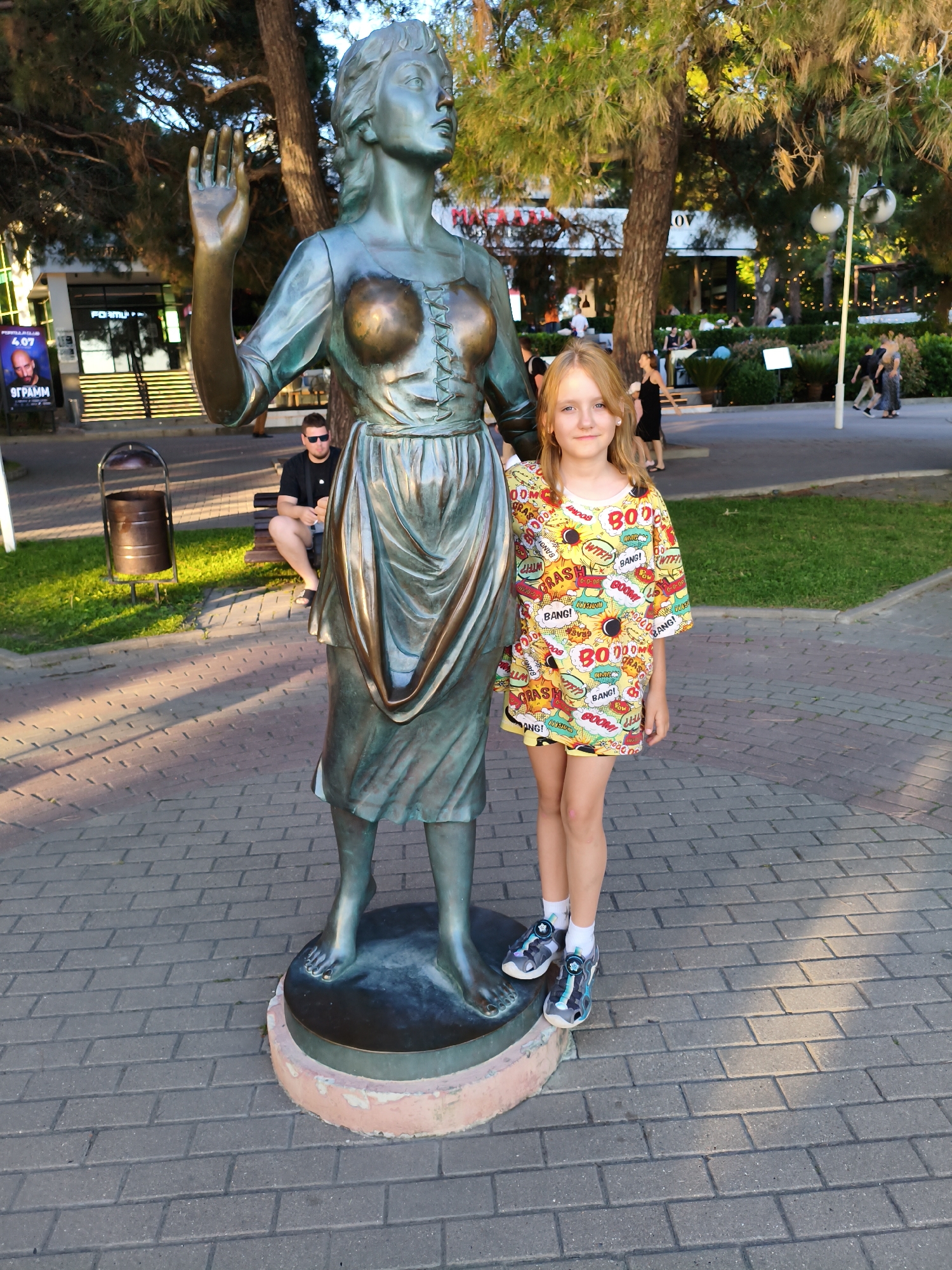 A young girl with blonde hair and a colorful t-shirt adorned with comic book-style sound effects like "BOOM," "CRASH," and "BANG!" stands next to a bronze statue of a woman with her arm raised. The statue has a draped dress and prominent chest. The girl is wearing white socks and sandals. In the background, a man in sunglasses sits on a bench, and a trash can is visible. Further back, there are trees, outdoor seating areas with people, and a building with a sign that has the Cyrillic text "FORMULA" and "ОГРАММ". The scene appears to be outdoors during the day, with sunlight casting shadows on the brick pathway. The atmosphere seems relaxed and casual, possibly a park or promenade.