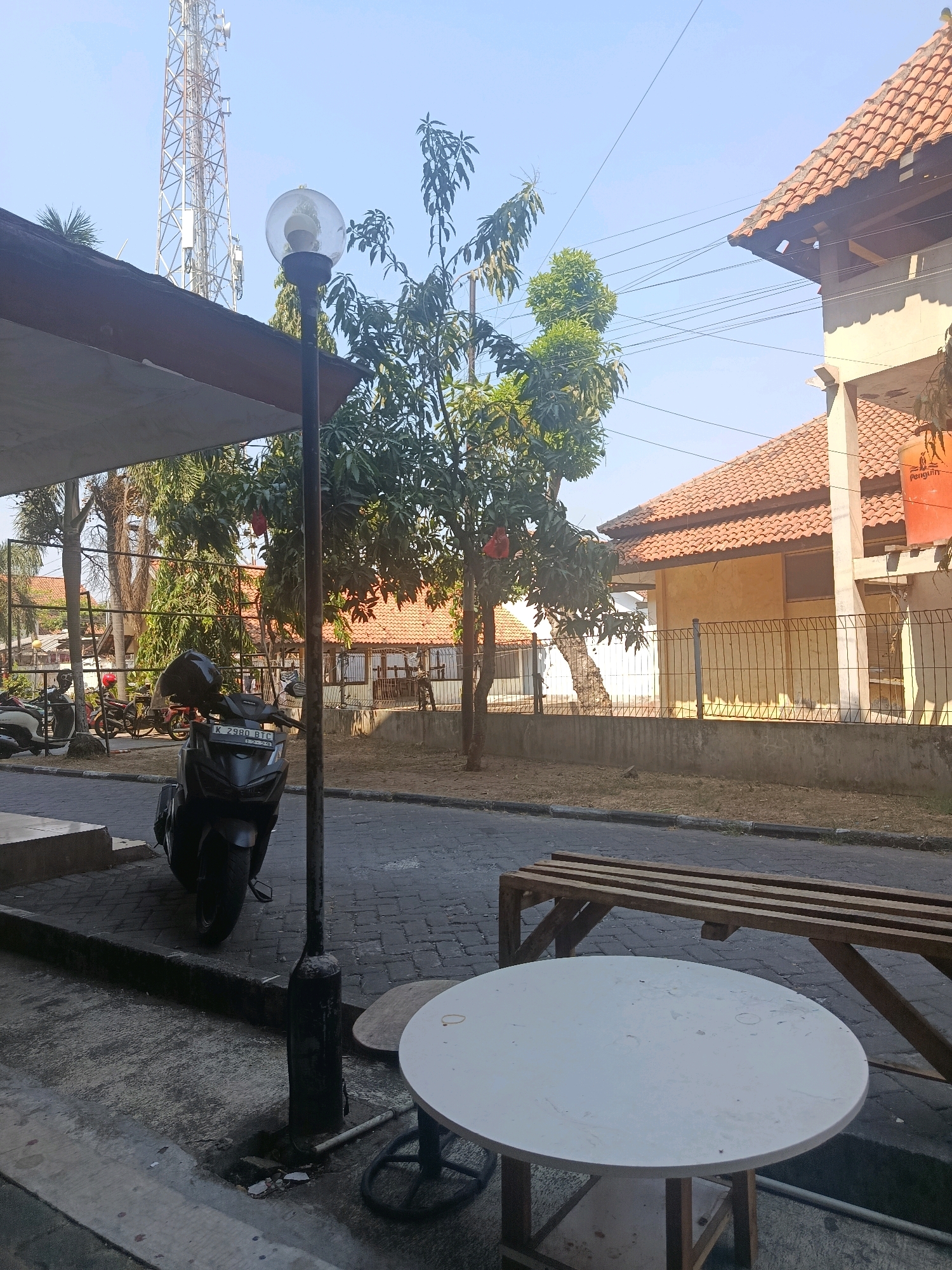 This is a street-level, medium-angle, full-color photograph taken outdoors on a sunny day. The scene depicts a street in what appears to be a residential or commercial area, possibly in Jobokuto, Indonesia. In the foreground, a white circular table with a wooden base is positioned on a paved surface. Behind the table, a long wooden bench made of planks is situated on a cobblestone street. To the left, a dark-colored scooter with a black helmet on its handlebars is parked by the curb. A black street lamp with a spherical light fixture stands near the scooter. Beyond the street, there are trees with green leaves and some red fruit. In the background, a two-story building with an orange tiled roof and yellow walls is visible, bordered by a metal fence. A tall cell tower can be seen in the distance. The sky is clear and blue.