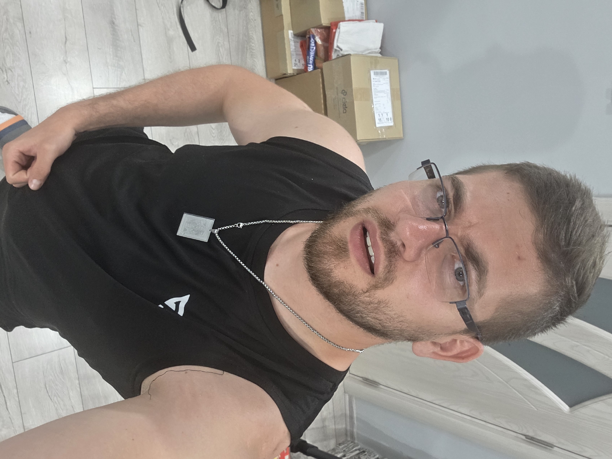 The image shows a young man, likely in his 20s or early 30s, taking an indoor selfie from a slightly upward, wide-angle perspective. The man has short brown hair, a beard, and wears dark-framed glasses. His mouth is slightly open, and his eyes are wide, conveying a surprised or engaged expression. He is wearing a black t-shirt featuring a white, stylized 'A' or triangle logo on the chest, and a silver chain necklace with a rectangular pendant. His right arm is bent at the elbow, with his hand clenched at hip level, and part of his left arm is visible on the bottom left. A striped orange and white sock is visible on his left foot.
The background features light grey wood-look laminate flooring and a plain light grey wall. Stacked against the wall are several cardboard boxes with various labels. Partially visible text on these labels includes "Konnecto" on a blue/white label, "ciela," and numbers such as "12", "21", "111", "15", and "100". A dark strap or cable lies on the floor. A white interior door or cabinet door is partially visible on the right side of the frame. The setting suggests a domestic space, potentially indicating recent deliveries or moving activity. The image is oriented 90 degrees clockwise.
