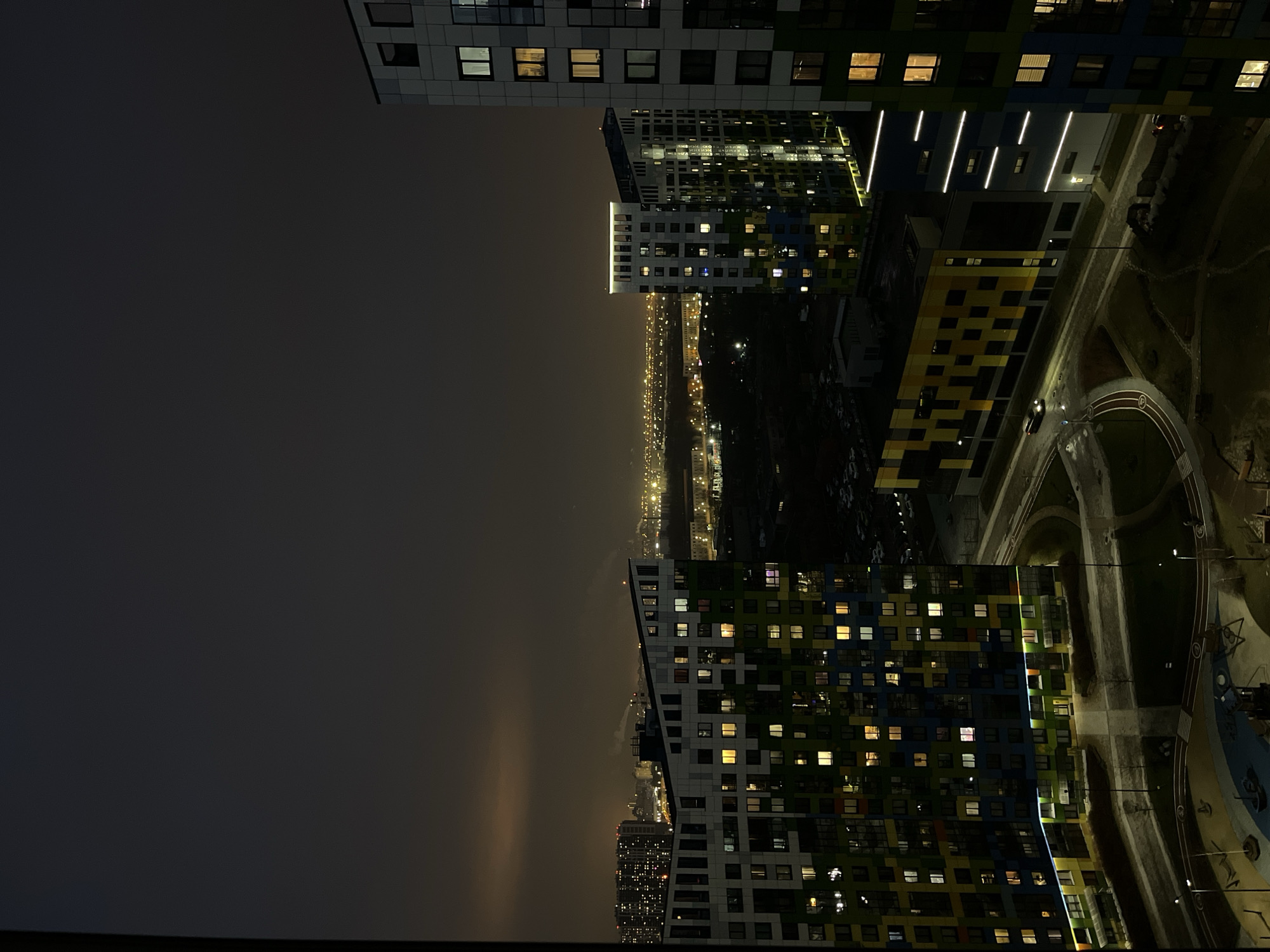The image depicts a nighttime urban landscape in Saint Petersburg, Russia. Several high-rise apartment buildings are visible, characterized by a modern architectural style with colorful facades. The buildings have numerous illuminated windows. A city skyline stretches in the distance, with lights indicating urban activity. A park and street area are visible below, with lighting illuminating pathways and roads. The sky is dark and overcast, with hints of light pollution. A road curves and streetlights are on. The orientation of the image is vertical.