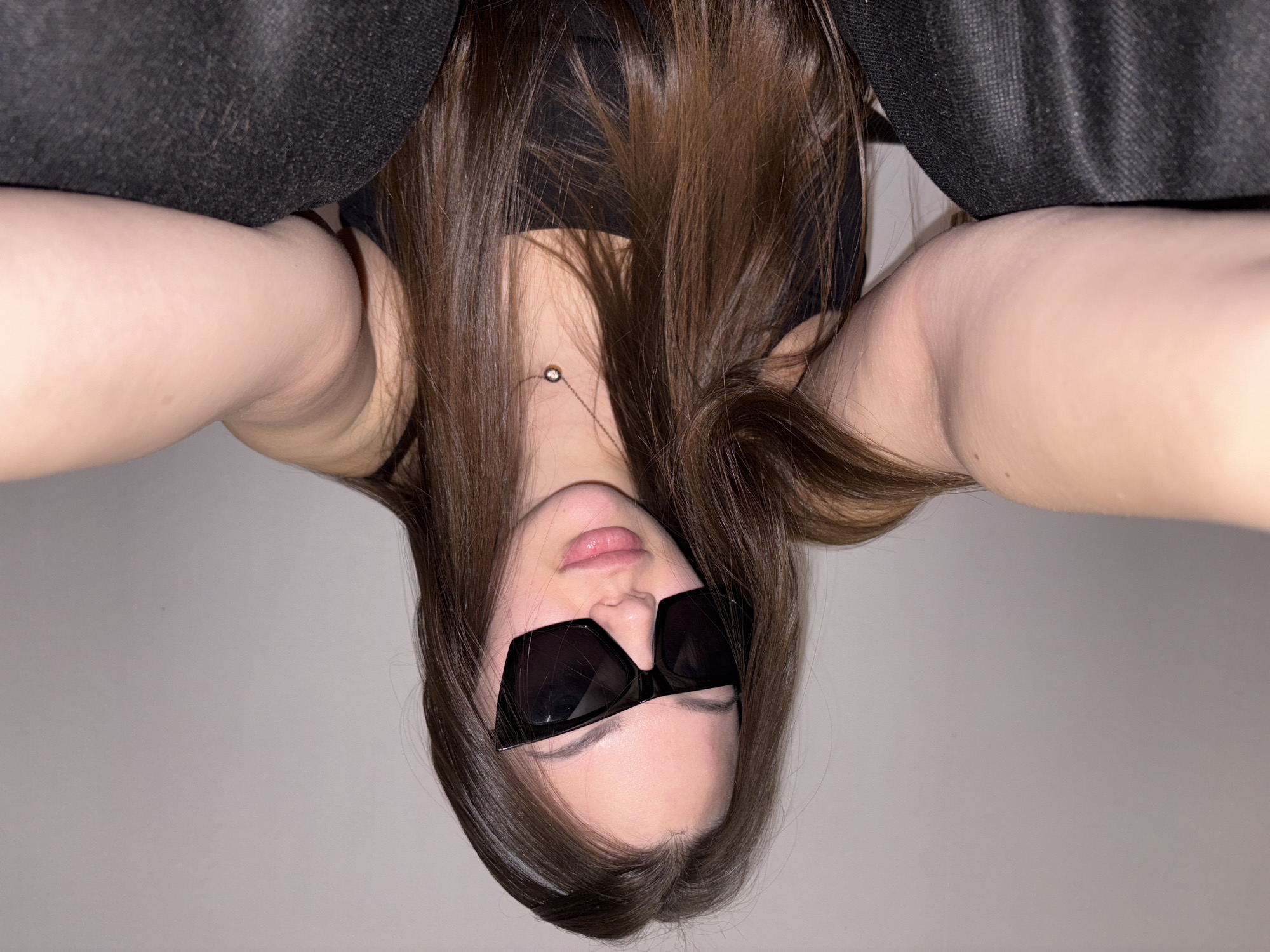 A selfie captures a young woman from a low-angle, upside-down perspective. She has long, brown hair that partially obscures a black tank top. She wears square, black sunglasses. Her skin is fair, and she has a simple necklace. The background consists of a plain, light-colored wall and a dark, textured fabric that could be clothing or furniture. The photograph gives no indication of the city of Chaikovsky, Russia.