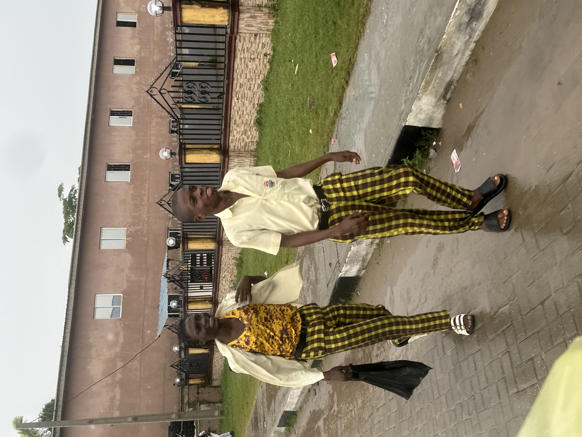 The image shows two young Black males standing outside a building. The building is a two-story structure with a brown, textured facade. It has several windows, some with white frames and others with darker frames. A black metal fence with decorative elements runs along the front of the building. The ground is wet, suggesting it has recently rained. There is a grassy area to the left of the building.
The subjects are both wearing plaid pants in yellow and brown. The person in the front is wearing a yellow and brown patterned tank top and a light-colored shirt over it, with the shirt open. They are holding a black umbrella. The person behind them is wearing a light yellow collared shirt with a badge on the left chest. Both individuals appear to be smiling or laughing. The setting appears to be a residential or institutional area, possibly a school, given the attire. The overall mood seems lighthearted and cheerful, despite the wet weather. There are no visible text elements in the image. The scene suggests an outdoor activity taking place possibly after or during a light rain shower.