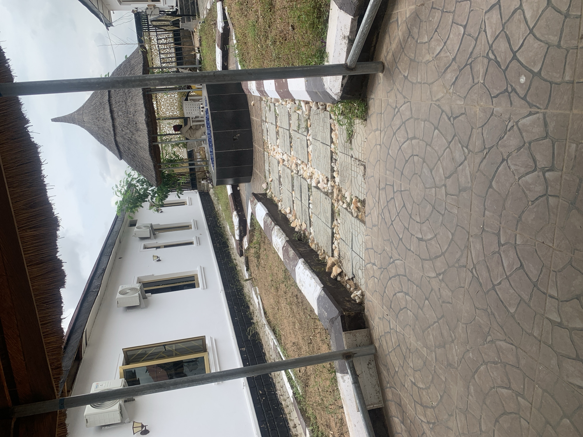 The image depicts an outdoor scene at a resort or residential compound in Jalingo, Nigeria, featuring a pathway leading towards a thatched gazebo. The pathway is constructed with stamped concrete tiles forming a circular pattern, interspersed with sections of square concrete pavers and gravel. A low, decorative border with alternating brown and white painted sections lines the pathway. To the left, a white building is visible, equipped with multiple air conditioning units and windows with gold-colored frames. To the right, beyond the pathway, there is a grassy area and a decorative metal fence. Inside the thatched gazebo, a person is seated at a table, partially visible. The overall environment suggests a tropical or sub-tropical climate, with a slightly overcast sky.