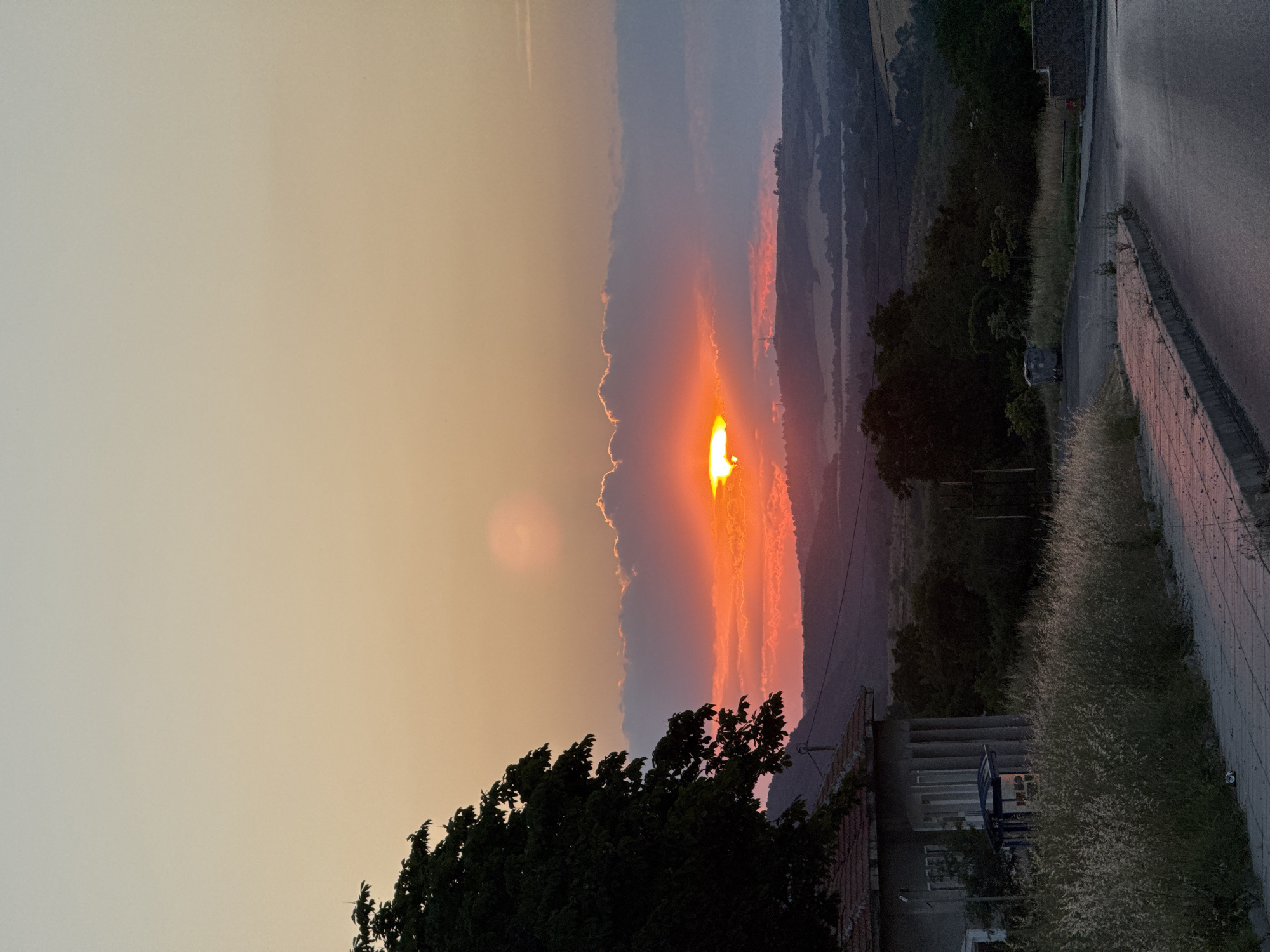 The image depicts a scenic sunset in Medovo, Bulgaria. The sun, a vibrant orange orb, is partially obscured by a dramatic cloud formation, casting warm hues of orange and pink across the sky. The sky transitions from a soft, pale yellow on the left to more intense sunset colors as it nears the horizon on the right. In the foreground, a large, dark green tree with dense foliage dominates the lower left portion of the frame.
The background reveals rolling hills and a valley, with layers of distant mountains fading into a dusky haze. A road with a paved sidewalk runs along the right side of the image, bordered by tall, dry grass. A few structures are visible, including a building with a tiled roof and a more modern building with a grey facade and what appears to be a blue bench or seating area in front of it. There are no people visible in the image. The time of day is clearly sunset, indicated by the colors of the sky and the low angle of the sun. The weather appears clear, with the clouds serving as a dramatic backdrop for the sunset. The overall emotion evoked is one of natural beauty and tranquility. No visible text is present.