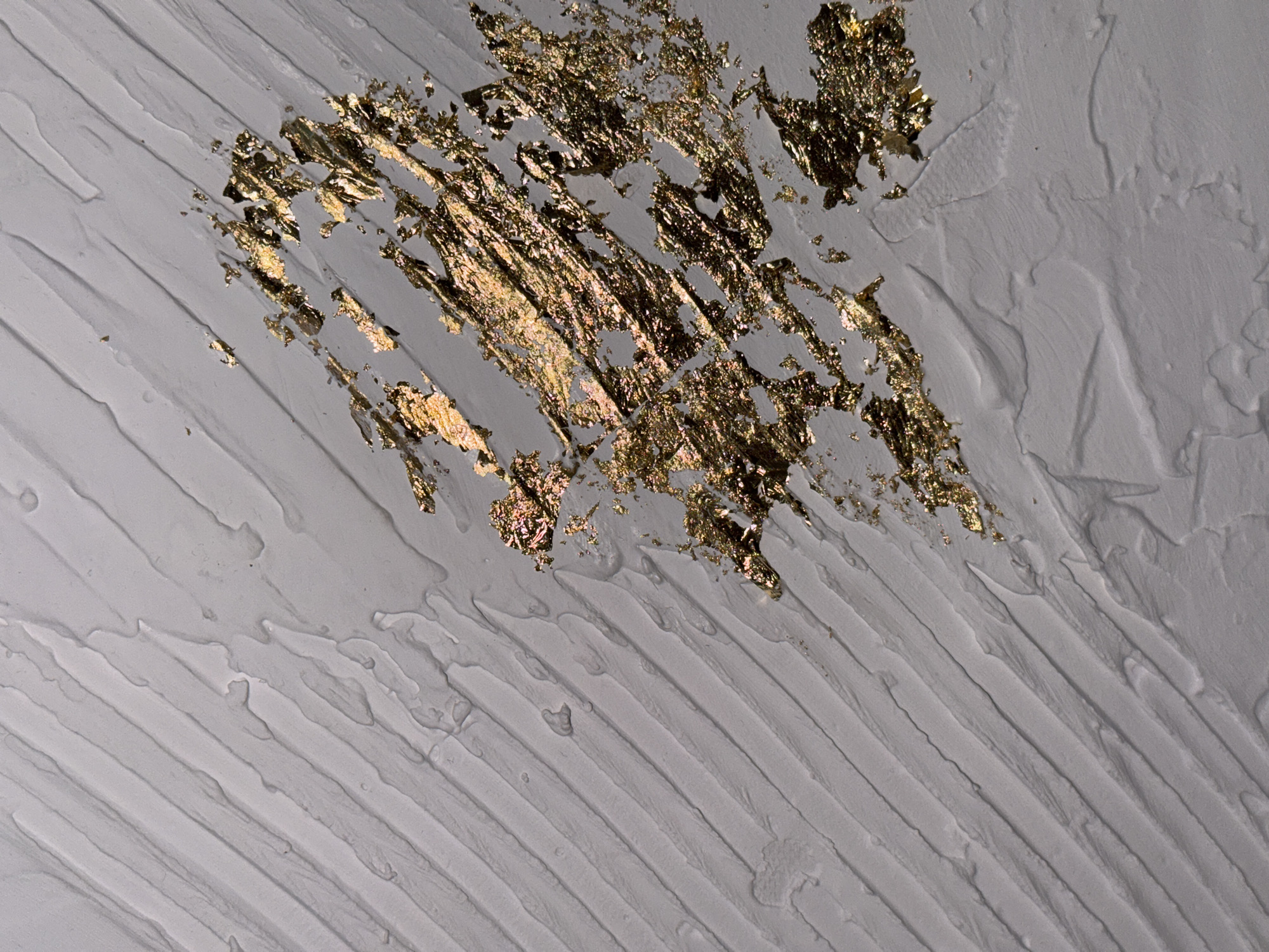 The image depicts a close-up of a textured surface, possibly a wall or artwork, featuring a gray or white base with visible, diagonally oriented brushstrokes or ridges. The surface is partially covered with fragmented, metallic, golden-colored flakes or leafing. The flakes are concentrated in a central area, thinning out towards the edges. The lighting is uneven, creating highlights and shadows that accentuate the texture of both the base surface and the flakes. The scene does not provide information suggesting it is located in Chaikovsky, Russia, or feature any people, subjects, objects, actions, or interactions beyond the described textures and materials.