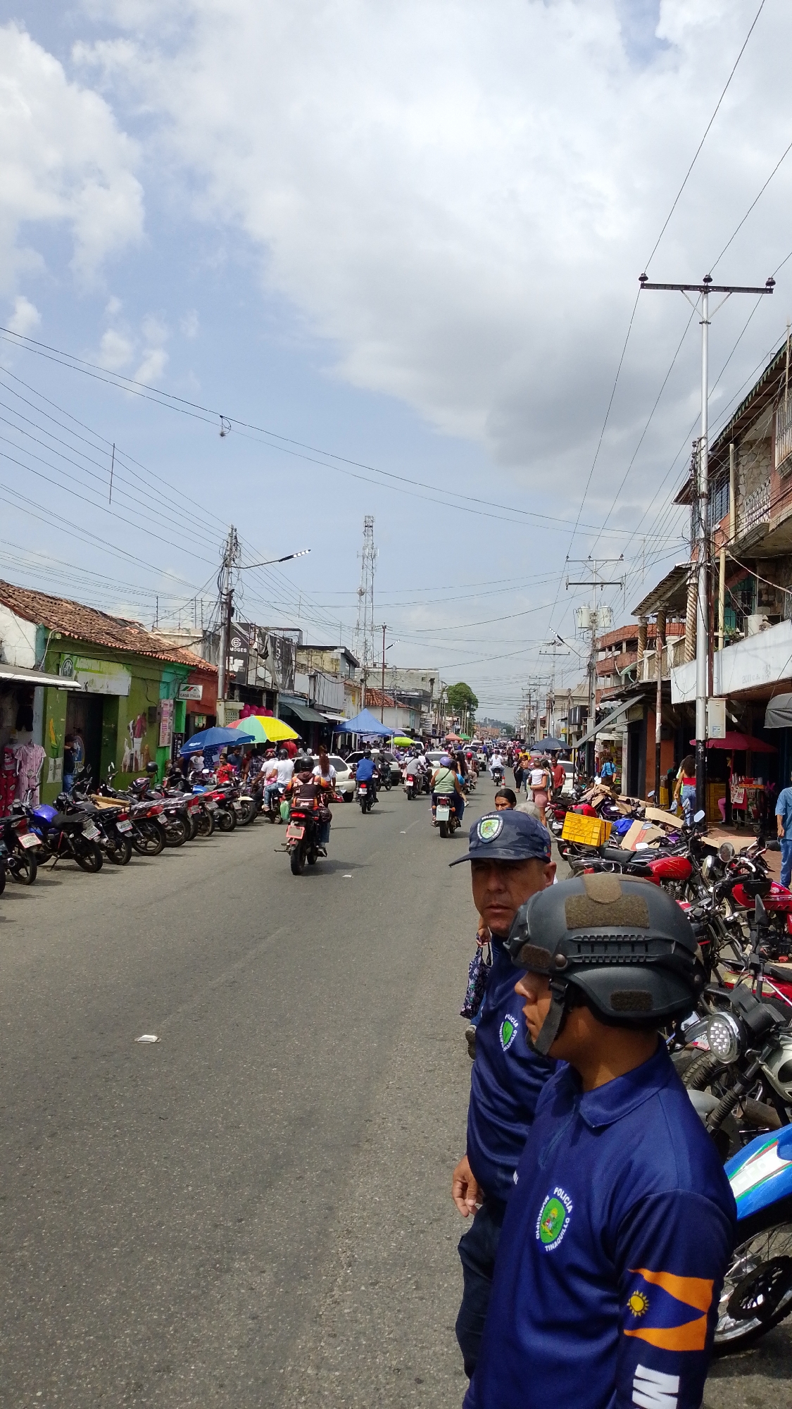 The image depicts a street scene in Tinaquillo, Venezuela. A street is filled with parked motorcycles and circulating motorcycle traffic. Two police officers stand in the foreground, wearing dark blue uniforms with the inscription "POLICIA MUNICIPIO TINAQUILLO" on their sleeves. One officer wears a peaked cap, while the other wears a tactical helmet. Behind them, the street is lined with buildings of varying heights and architectural styles. A mix of shops and residences can be observed. People are visible walking and gathering near the buildings. An assortment of vehicles including motorcycles and at least one white car navigate the street. Overhead, electrical and communications wires crisscross the sky. In the distance, a telecommunications tower is visible. The sky is partly cloudy.