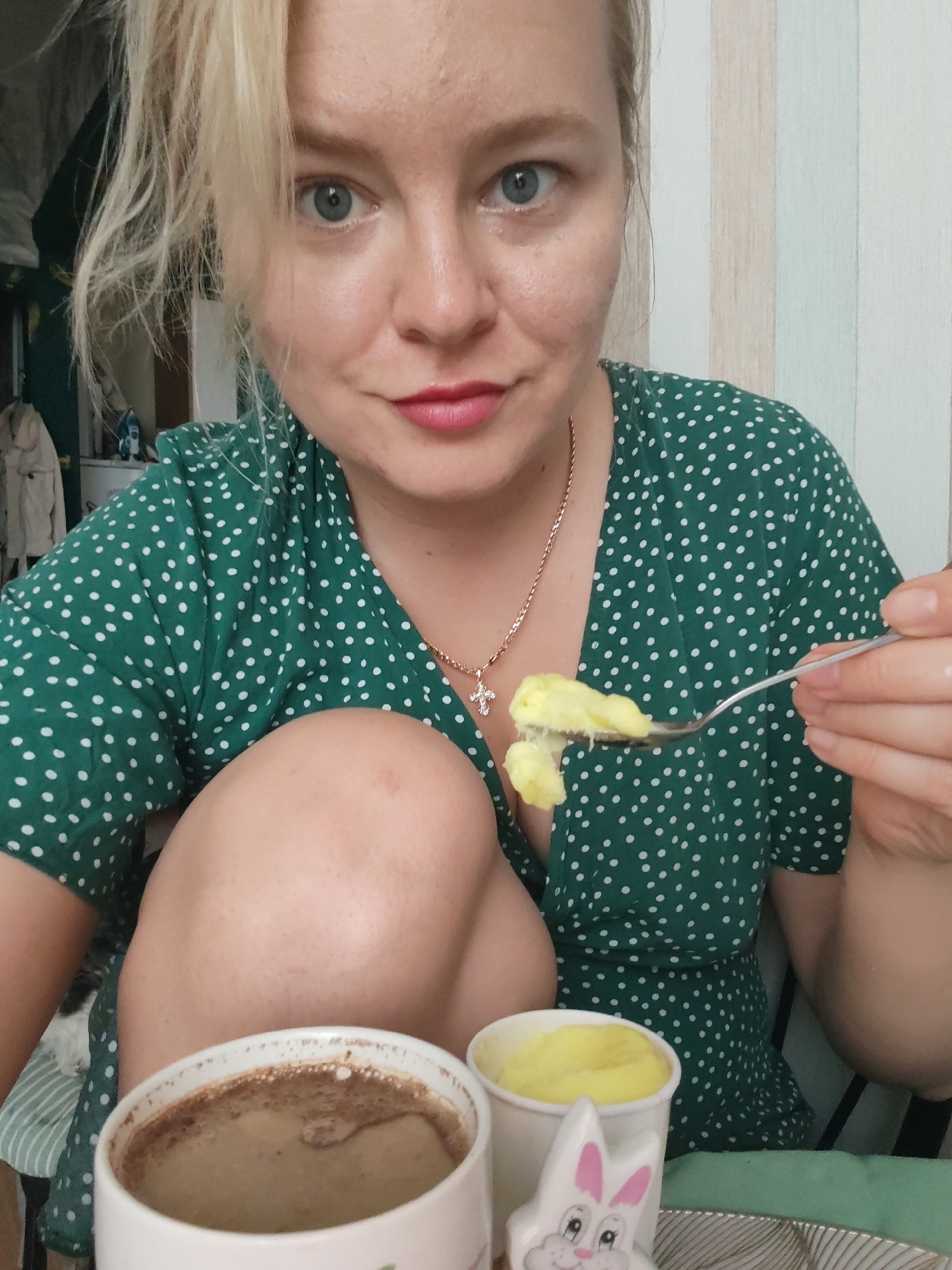 In Izhevsk, Russia, a close-up, eye-level shot shows a woman with blonde hair and blue eyes eating. She is wearing a green polka dot dress and a gold necklace with a cross pendant. In her right hand, she holds a fork with yellow food. In front of her are a white mug of coffee and a small paper cup filled with yellow food. A white ceramic bunny decoration is also visible. The background features a wall with vertical stripes.