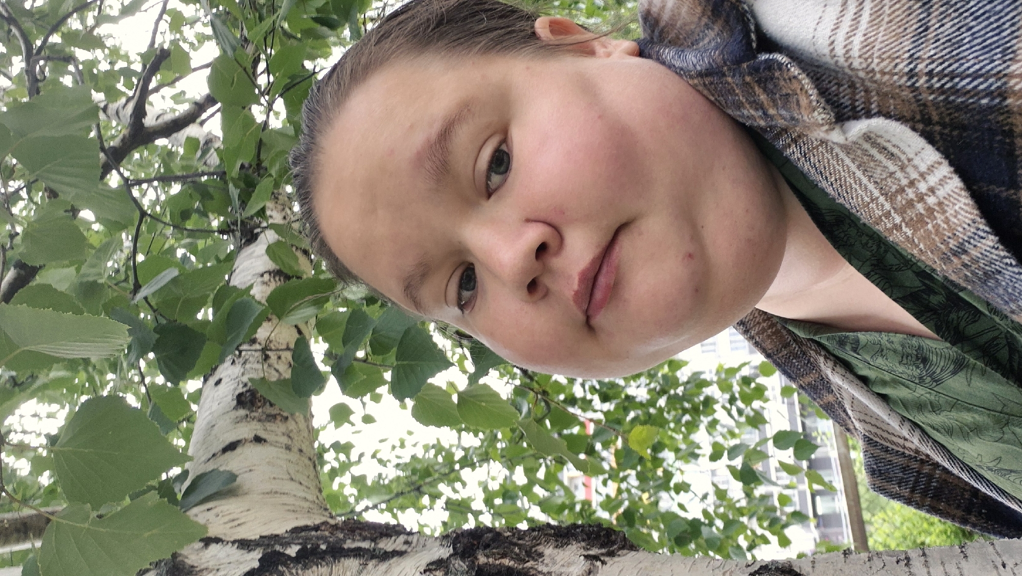 A medium shot captures a woman from a low angle, positioned near a birch tree. The tree's white bark with black markings is visible in the foreground, with green leaves filling the background. The woman, wearing a plaid jacket over a green patterned shirt, looks down at the camera. In the background, a multi-story building is partially visible. The location is Chaikovsky, Russia.