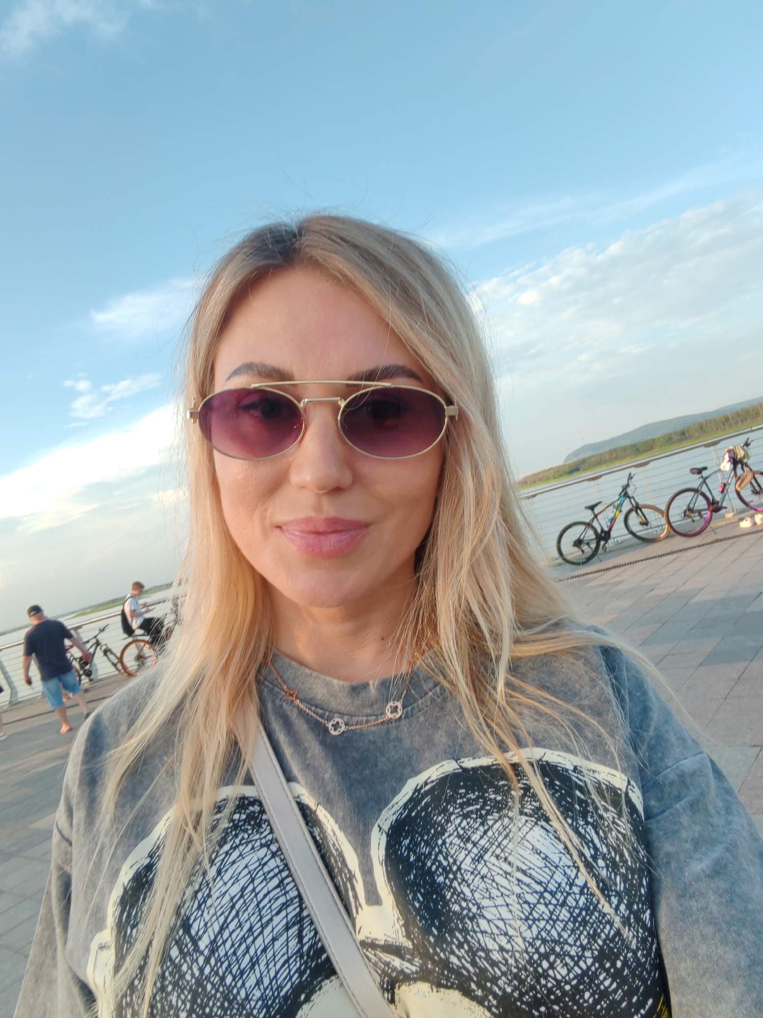 A woman with blonde hair, wearing sunglasses and a gray sweatshirt with a graphic print, poses for a selfie outdoors. She has a light-colored purse strap across her chest and a necklace. In the background, on a paved walkway next to a body of water, several bicycles are parked. Two men are visible in the middle ground: one is walking away from the camera wearing a dark t-shirt and blue shorts, and another is riding a bicycle. The scene appears to be in Komsomolsk-on-Amur, Russia, as evidenced by the surrounding landscape which includes a river and distant hills. The sky is blue with scattered clouds.