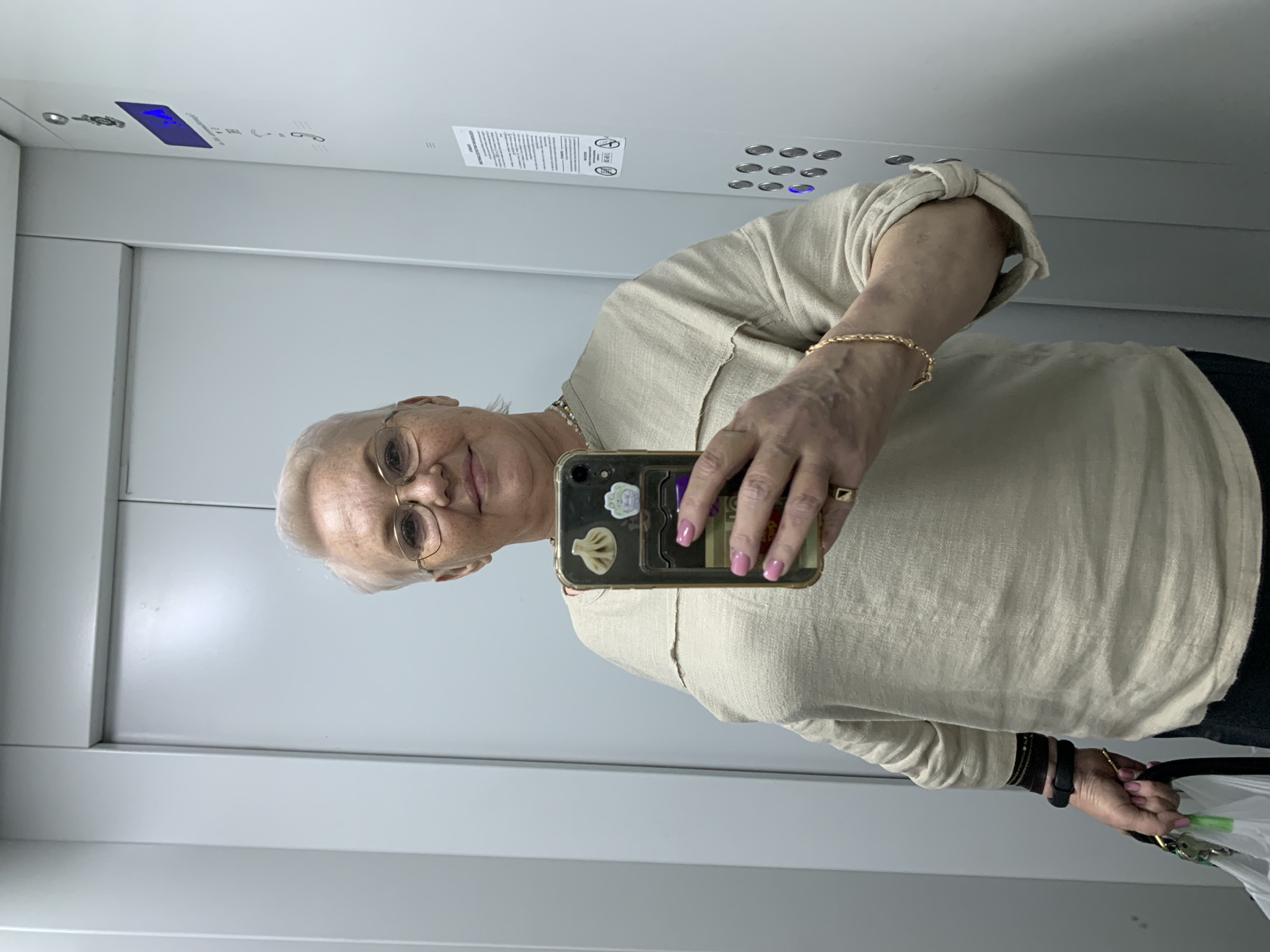 A fair-skinned woman with short gray hair and glasses is taking a selfie in what appears to be an elevator. She is wearing a light-colored blouse, and a gold bracelet. Her fingernails are painted pink. She is holding a smartphone with stickers on the back in her right hand. A gold ring is visible on her ring finger. She is holding a white plastic bag with green objects inside in her left hand. The elevator has silver walls, a panel of buttons, and a digital display at the top. A sign is posted on the elevator wall near the buttons. Based on the metadata, the location is Chaikovsky, Russia.