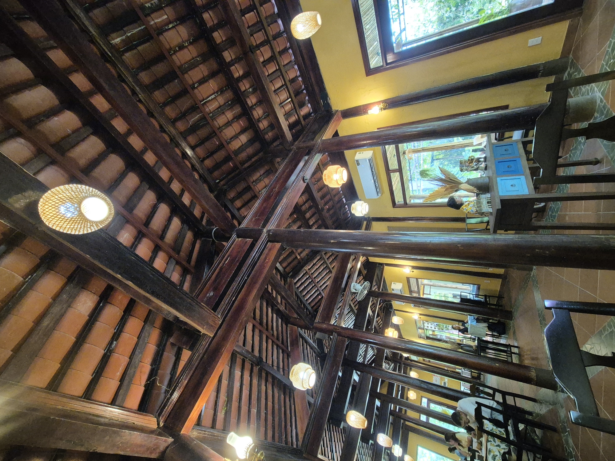The image depicts the interior of a restaurant in Da Nang, Vietnam, characterized by traditional Vietnamese architecture. The ceiling is constructed with dark wooden beams and covered with orange terracotta tiles. Several woven pendant lights with warm illumination are suspended from the beams. Large, dark wooden pillars support the roof structure, creating a rustic and natural ambiance.
The walls are painted a vibrant yellow, contrasting with the wooden elements. Large windows line the walls, offering views of lush greenery outside, suggesting the restaurant is situated in a garden or natural setting.
In the foreground, dark wooden chairs and tables are visible, indicating a dining area. The floor is tiled with terracotta and some mosaic patterns.
In the middle ground, patrons are seated at tables, engaged in dining or conversation. One person is visible at a table near a window, examining something on the table. Further back, another individual is seated at a table by a window, looking out.
A notable feature in the mid-ground is a blue cabinet with drawers, set against the yellow wall. On top of it, decorative items are placed, including a vase with dried foliage and figurines. An air conditioning unit is mounted on the wall above a window.
The overall impression is one of a serene and aesthetically pleasing dining environment, blending traditional Vietnamese design with natural elements.