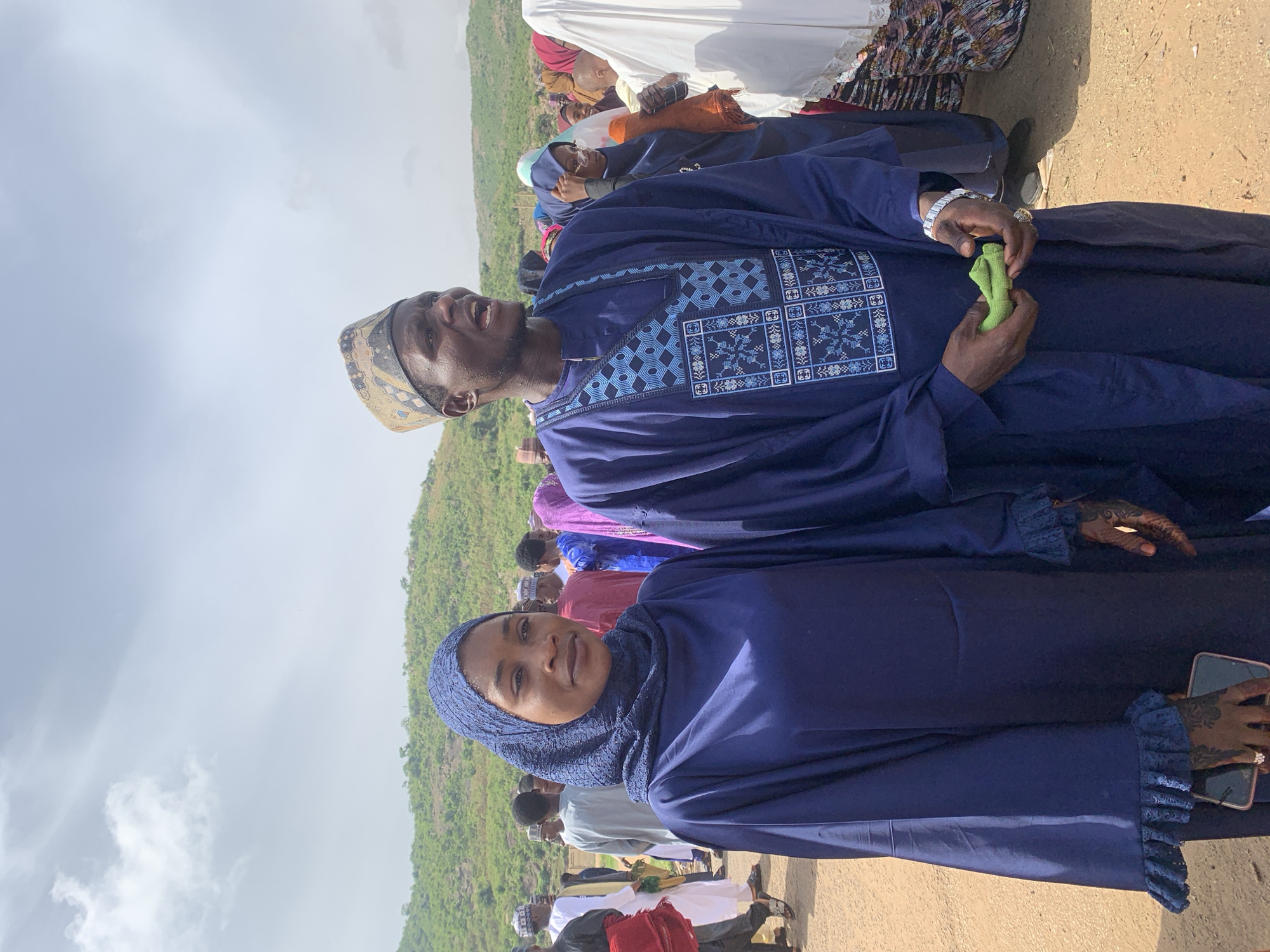 In Naraguta Hausawa, Nigeria, a man and a woman are prominently featured in an outdoor gathering. The man, wearing a blue traditional robe with intricate embroidery on the chest, also sports a patterned cap and a silver watch. He holds a small green object in his hands. Beside him, the woman is dressed in a matching blue hijab and robe, adorned with ruffled cuffs. She holds a mobile phone in her right hand, and both hands display henna designs. They are part of a larger group of people, visible in the background, some wearing hijabs and traditional attire. The terrain appears to be a dirt ground with a lush green hill in the distance under a cloudy sky.