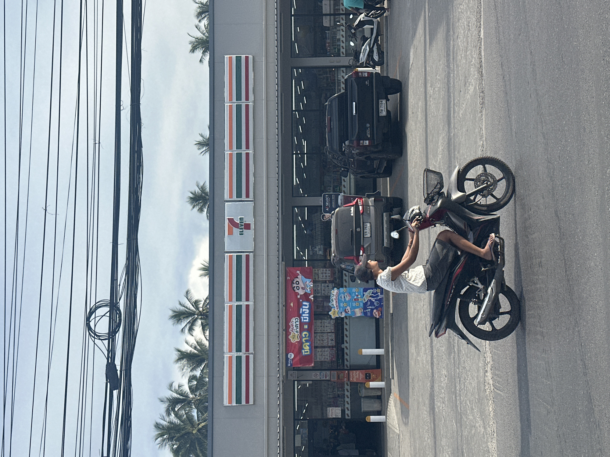A person rides a motorcycle in front of a 7-Eleven store in Baan Mae Nam, Thailand. The 7-Eleven store has its signature red, white, and green striped logo above the entrance, and several displays and advertisements are visible inside the store through the large glass windows. Outside the store, there are parked vehicles, including a black pickup truck and a scooter. Palm trees are visible above the store's roofline, and a tangle of power lines is strung across the sky. The rider is wearing a light-colored patterned shirt and dark shorts, and is seen from the side as they move across the paved road.