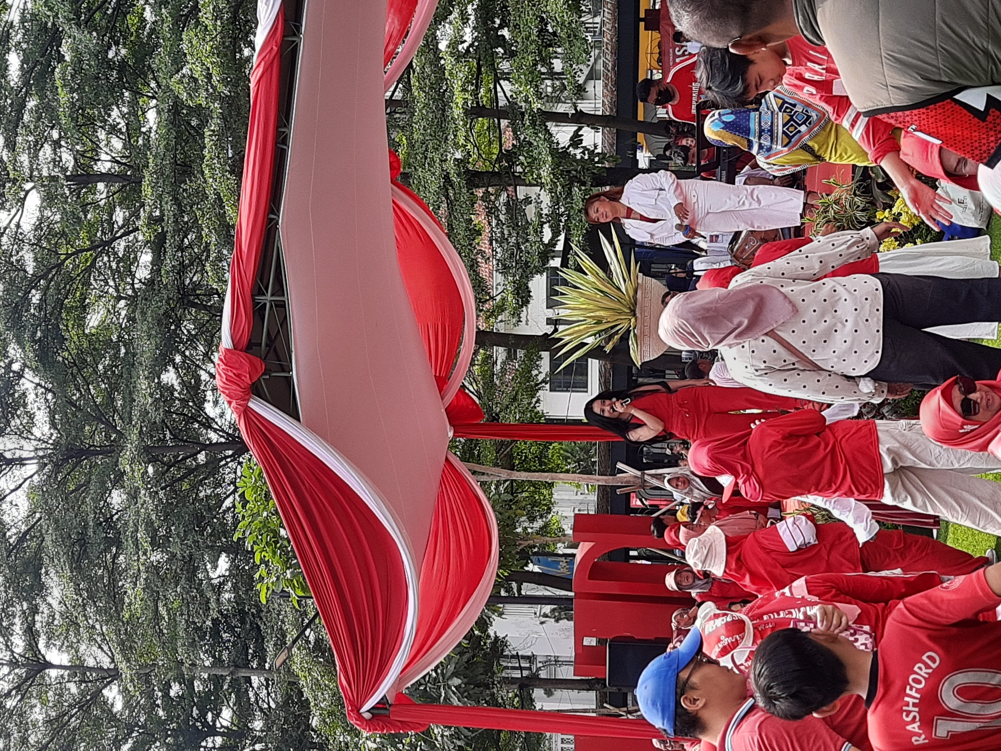 The image depicts an outdoor event with a stage draped in red and white fabric, reminiscent of the Indonesian flag. Two women are on the stage, one in a red jumpsuit singing into a microphone, and another in a white jacket and pants also holding a microphone. A crowd of people, many wearing red attire, are gathered in front of the stage, suggesting a celebratory or nationalistic event. Large red letters are visible behind the performers. In the foreground, a young boy in a red shirt with "Rashford 10" printed on it is looking towards the stage. The background features lush green trees and buildings, indicative of an urban park or public square. The presence of the Indonesian flag colors and the gathering of people suggest a public celebration or festival, possibly in Bandung City, Indonesia, given the context.