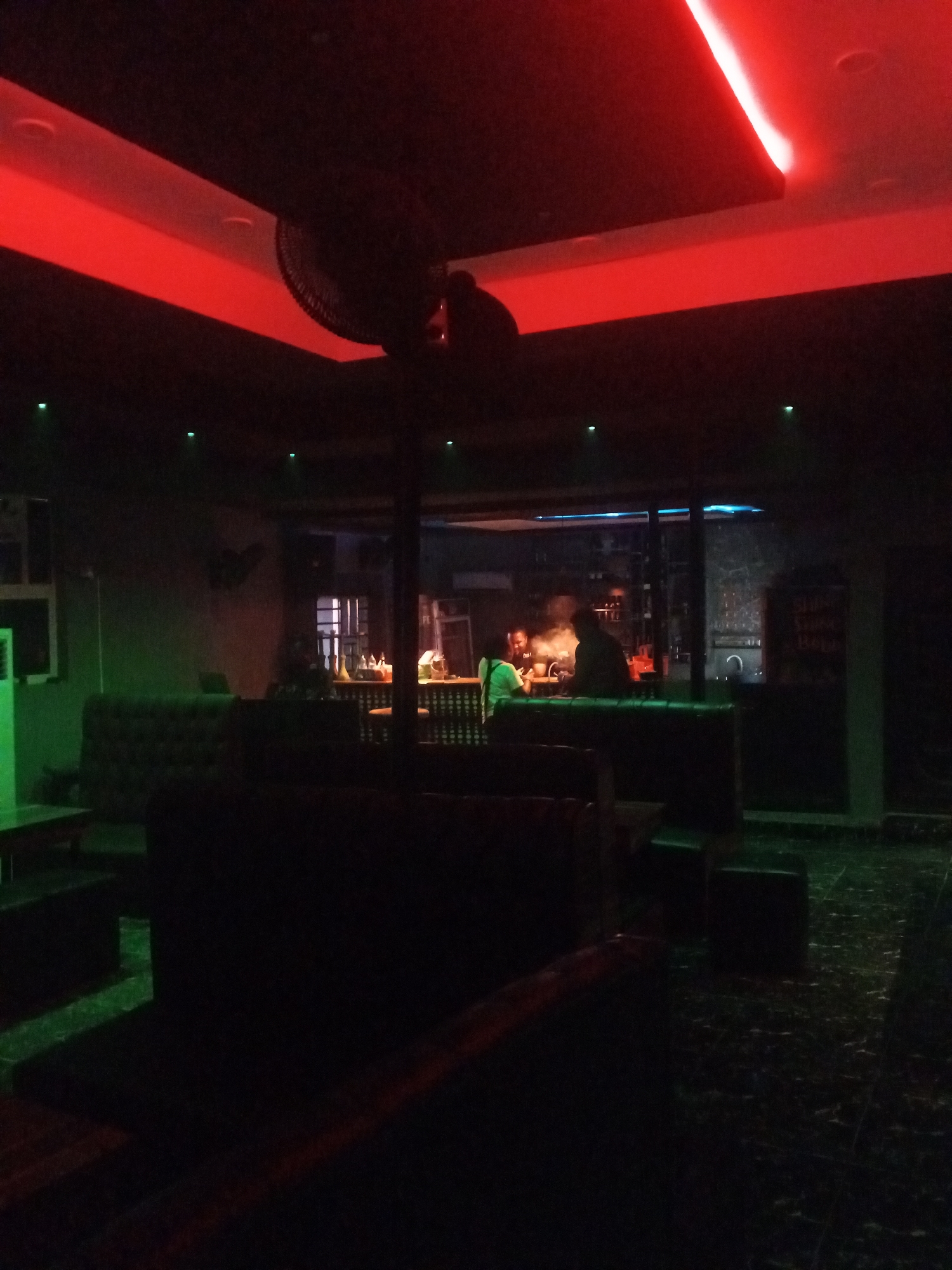 This media file captures the dark interior of what appears to be a bar or lounge in Lokoja, Nigeria, illuminated by vibrant artificial lighting, suggesting it is nighttime or evening. Red LED strip lights line the ceiling, casting a strong red glow across the upper part of the room and parts of the wall. Green spotlights or downlights are strategically placed, illuminating sections of the floor and an air conditioning unit on the left wall.
The scene features comfortable-looking, dark upholstered sofas or booths arranged in the foreground and midground, implying a relaxed seating arrangement for patrons. A ceiling fan, largely in silhouette, is visible in the upper center, partially obscuring the red ceiling light.
In the background, a brightly lit bar counter serves as a focal point. Two individuals are visible near the bar; one person wearing a light green shirt is seen from behind, while another figure is a darker silhouette, possibly a bartender or another patron. Bottles and other bar equipment can be faintly discerned on the counter, suggesting drink preparation or service.
To the right of the bar, a dark sign or chalkboard is partially visible, with some illegible text. However, the word "SHISHA" can be made out near the top of the sign, indicating that shisha is likely offered at this establishment. The overall ambiance is dim and intimate, characteristic of a nightspot designed for socializing and leisure.