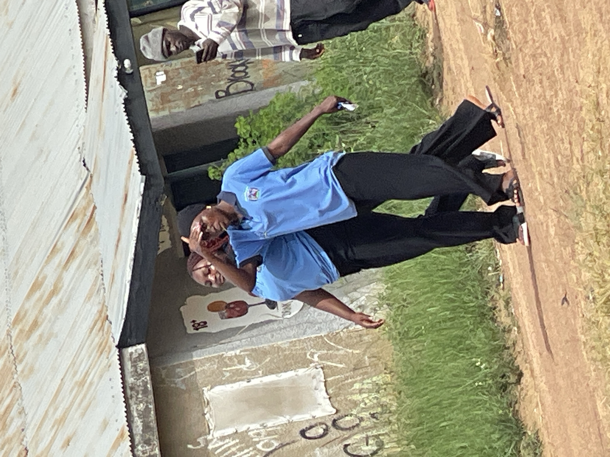 In Durumi, Nigeria, three individuals are captured in an outdoor scene. Two young women, dressed in matching bright blue short-sleeved polo shirts and dark trousers, walk side-by-side. The woman on the left has her left hand raised to shield her eyes from the sun. The woman on the right wears a black headscarf and is also wearing a blue polo shirt and dark trousers. She holds a small white object in her right hand. A third individual, a man wearing a grey beanie, a horizontally striped shirt, and dark trousers, stands to the right, looking down at a mobile phone in his hand. The background features a weathered building with a corrugated metal roof and graffiti on its exterior walls. Green foliage and a dirt path are visible in the foreground, suggesting a somewhat rural or unpaved urban setting. The overall lighting indicates a bright, daytime environment.
